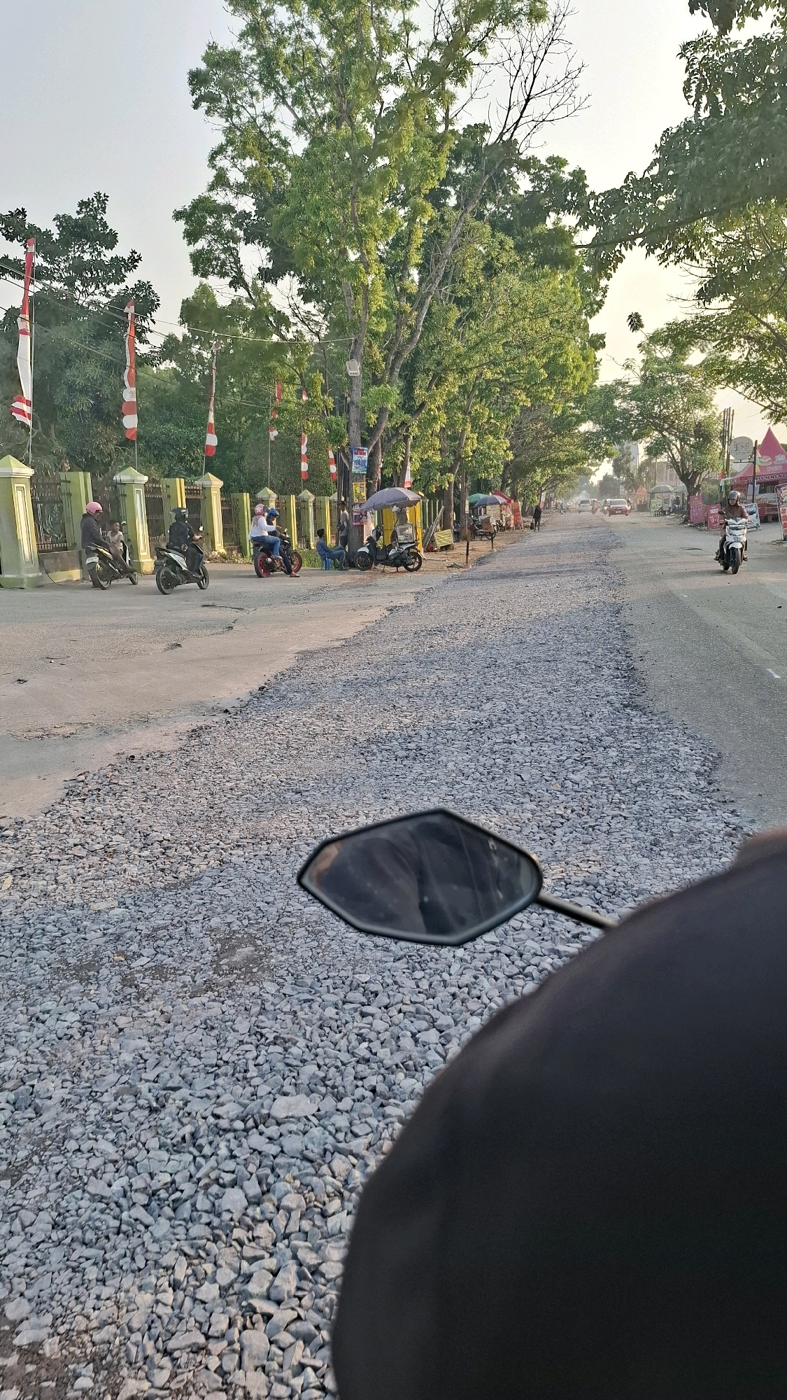 The image captures a street scene in Bandar Lampung, Indonesia, possibly during daylight hours given the lighting conditions. The road ahead is partially paved, with a significant portion covered in gravel, suggesting ongoing construction or repair work. Several trees line the left side of the road, their leaves green, indicating a temperate or tropical climate.
In the foreground, the handlebars and a portion of the mirror of a motorcycle are visible, indicating that the photograph was taken from the perspective of a rider. Further down the road, several people are seen on motorcycles, some appearing to be stopped or moving slowly. Red and white flags, possibly Indonesian flags, are displayed on poles along the left side of the street, attached to a green fence.
Various roadside stalls or temporary structures are present on the right side of the road, with some displaying umbrellas for shade. A few cars and motorcycles are visible further along the street, suggesting moderate traffic. The overall impression is a street in a developing urban area, with elements of commerce and infrastructure development coexisting.