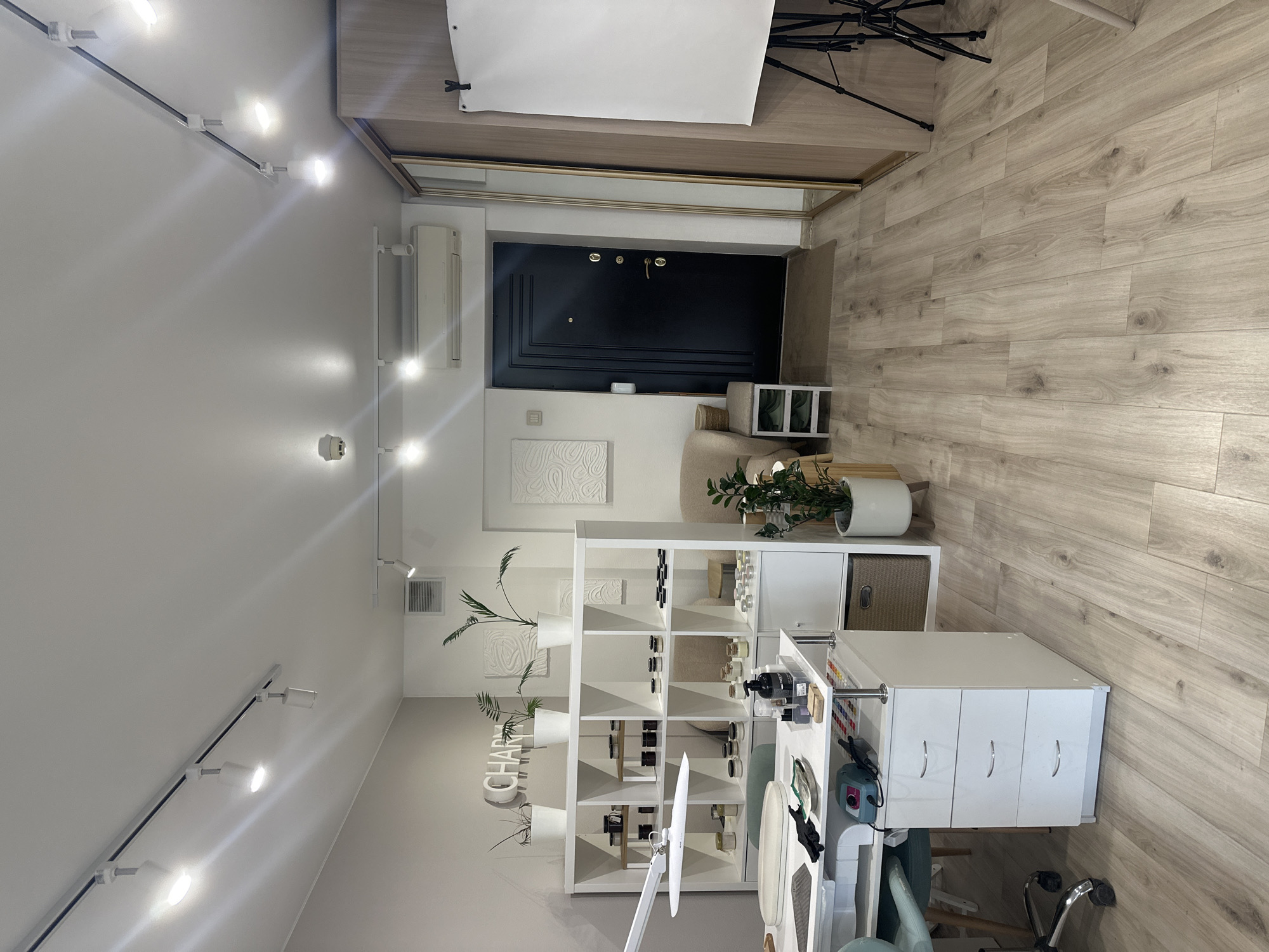 The image shows an interior view of a room, possibly a salon or studio, in Perm, Russia. The room is well-lit with track lighting. Predominantly white walls and light wood flooring contribute to a clean and modern aesthetic. Key objects include a black door, white shelving units with various items displayed on them (possibly beauty products or salon supplies), plants in white pots, and a white desk or workstation with drawers and equipment. A chair is positioned near the desk. The room has minimalist decor, including textured white artwork on the walls. A clothing rack and a photography umbrella reflector are visible near the entrance. There are no people visible in the shot, implying a static scene ready for or recently vacated.