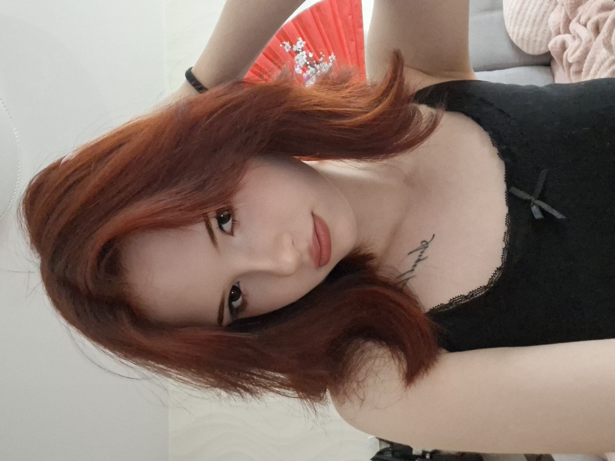A young woman with red hair is lying down, facing the camera. She is wearing a black top with a lace trim and a small bow. There is a red fan in the background. She has a tattoo on her chest. The environment appears to be indoors. There is no discernible information indicating this image was taken in Chaikovsky, Russia.