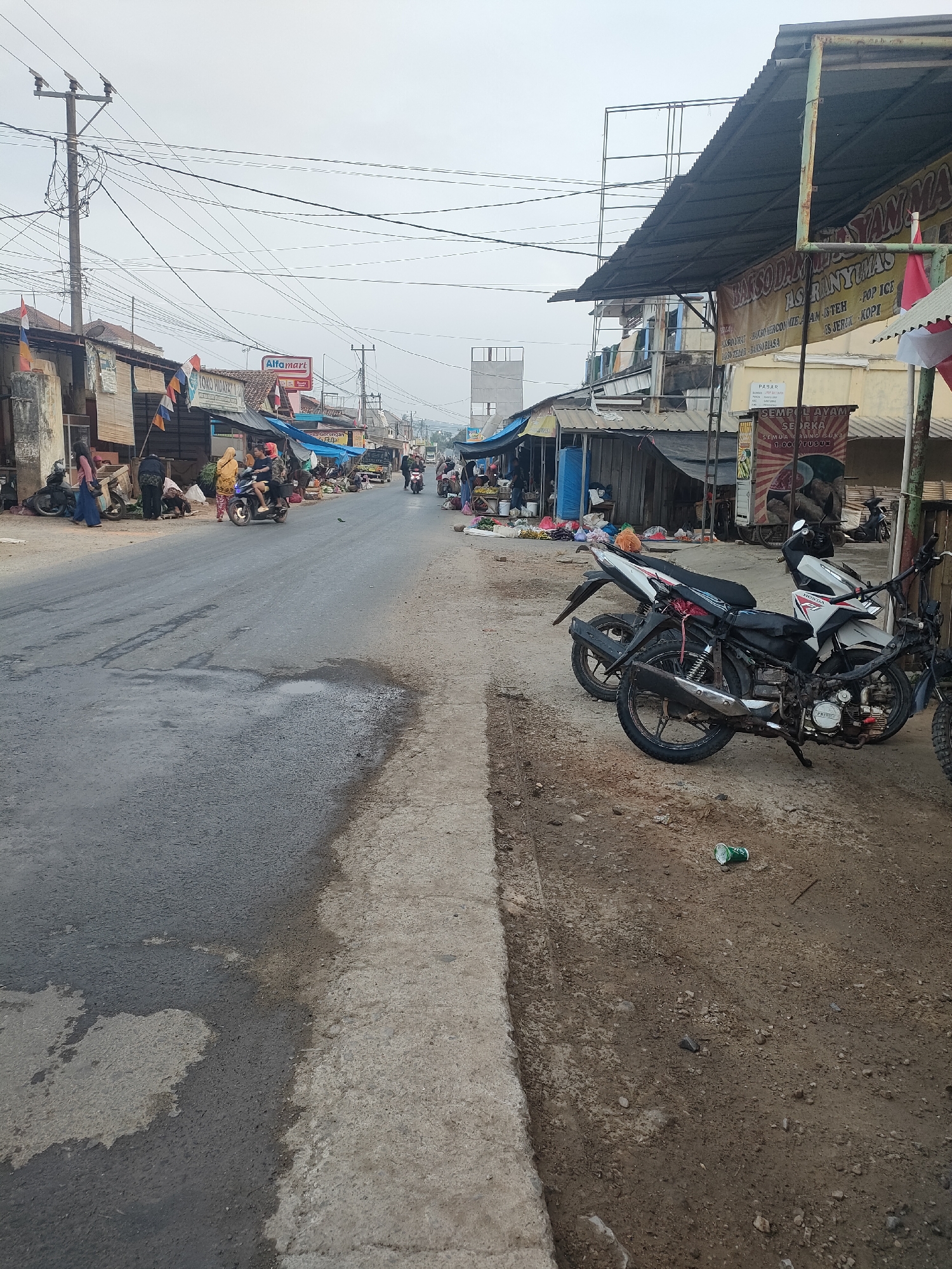 This is a ground-level, eye-level, medium shot of a street scene in Sukoyoso, Indonesia. The street is paved with asphalt and shows signs of wear, with patches of water on the surface. To the right, a dirt and gravel shoulder runs alongside the road, littered with debris including a crushed green can and a rusty metal rod.
Along the left side of the street, a row of market stalls and shops are visible, some with corrugated metal roofs. A prominent sign for "Alfamart" is displayed. People are interacting at the stalls, with some individuals appearing to be vendors and others customers. Several motorcycles are parked or in motion.
On the right side of the street, several motorcycles are parked along the dirt shoulder. The motorcycles are of varying models, with one particularly visible in the foreground. The background shows a mix of buildings and utility poles with a complex network of overhead wires. The sky is overcast, indicating a potentially cloudy or hazy day.