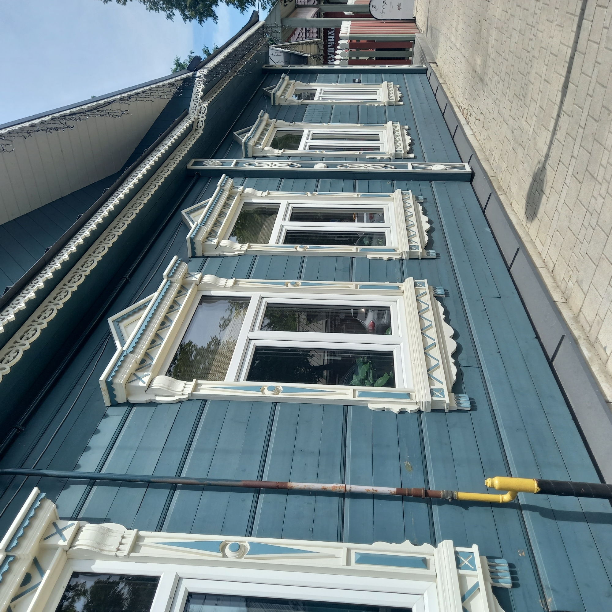 The image displays the exterior side of a two-story wooden building located in Zvenigorod, Russia, under a bright, sunny sky. The building's facade is painted a deep teal or blue-green, constructed from horizontal wooden planks. Five white-framed windows are visible, each featuring elaborate decorative wooden trim, known as nalichniki, painted in white and a lighter shade of teal-blue. These trimmings incorporate various patterns, including scalloped edges, geometric triangles, "X" motifs, and vertical elements with circular and pointed inserts.
Above the windows, along the roofline, an intricate white carved wooden frieze with repetitive curvilinear and floral patterns adorns the eaves. From the underside of these eaves, multiple dark, thin strings or chains hang vertically. A dark brown and white section of what appears to be a drainage or utility pipe runs vertically along the wall between the first and second windows from the left, transitioning to a yellow and black painted section towards the bottom.
The ground directly in front of the building is paved with light grey rectangular interlocking bricks. A dark grey base or foundation separates the wooden siding from the pavement. Reflections of green trees, a light blue sky, and a white car are visible in the windowpanes. In the background, further down the street to the right, other buildings with dark roofs and a wooden fence are partially discernible, along with a white sign bearing dark text. No individuals are present in the scene.