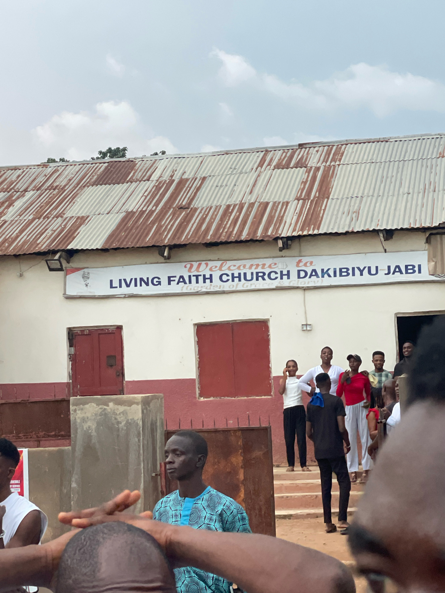 The image captures an outdoor scene in front of a building with a sign that reads "Welcome to LIVING FAITH CHURCH DAKIBIYU-JABI (Garden of Grace & Glory)". The building has a weathered, corrugated metal roof and a white facade with red trim around the windows and doors. Several people are visible in the foreground and on steps leading up to the building. Some individuals are looking towards the camera, while others appear to be in conversation. The environment suggests a community gathering or event taking place at the church. The presence of the church's name, "DAKIBIYU-JABI," strongly indicates the location is in Abuja, Nigeria, as this is a known area within the city.