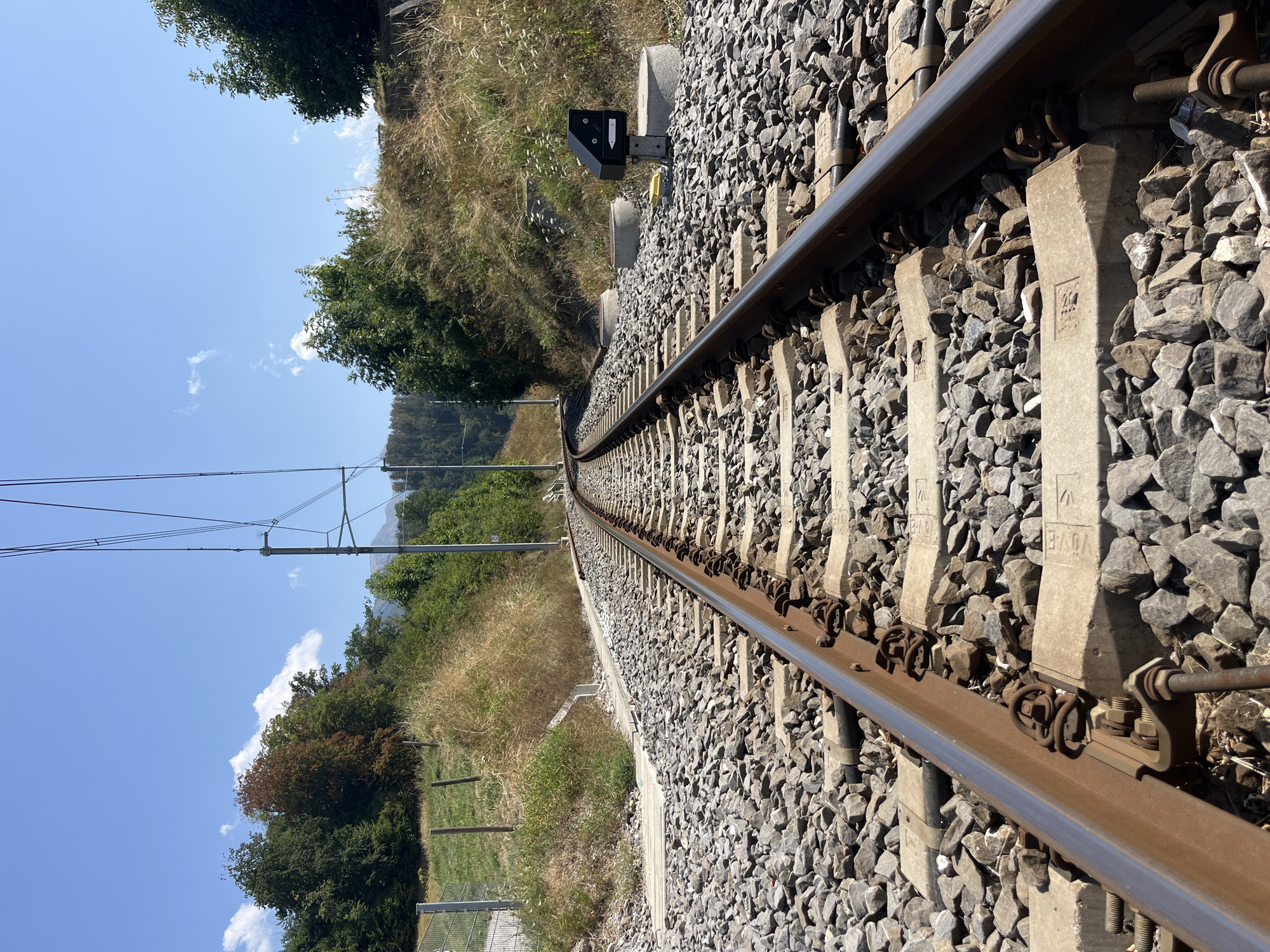 The image shows a set of railroad tracks curving to the left, surrounded by a ballast of grey stones. The tracks are made of metal rails secured to concrete sleepers. On the right side of the tracks, there is a small black box with a white arrow pointing left, mounted on a pole and secured to a concrete base. Beyond the tracks, the terrain slopes upwards, covered with dry grass and scattered green trees. Further in the background, more trees and a distant mountain range are visible under a clear blue sky with a few scattered clouds. The scene appears to be in an outdoor, rural setting, likely in Pringy, Switzerland, given the context. There are no people or any ongoing actions depicted in the image.