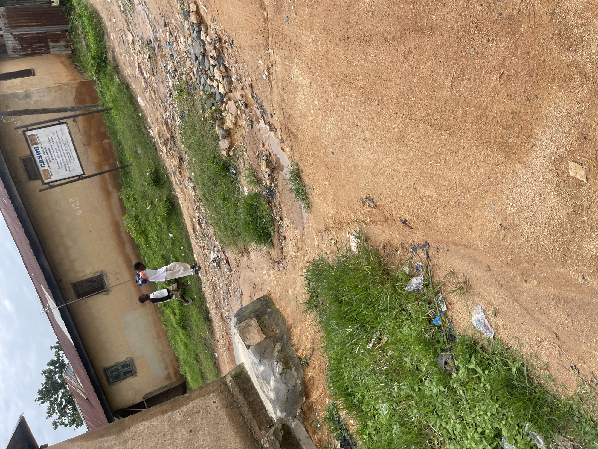 The image depicts an outdoor scene, likely in Jos, Nigeria, characterized by a rustic and somewhat neglected environment. In the background, a light-colored building with a corrugated metal roof stands, showing signs of age. A sign on the building, partially visible, reads "CASOR", with additional text below it detailing schedules for various activities. To the left of the building, two young boys are walking. The boy in front wears a white shirt and light-colored pants, while the boy behind him is in a black and white shirt and shorts. The ground in the foreground is a mix of reddish-brown dirt and gravel, with patches of green grass and scattered debris, including plastic bags. There are puddles of water on the ground, suggesting recent rain. The overall impression is of a common, everyday setting in a residential or community area.