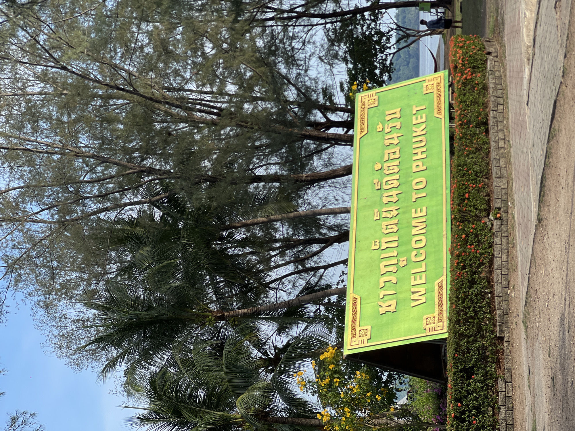 The image shows a "Welcome to Phuket" sign in Phuket City Municipality, Thailand. The sign is green with gold lettering and is positioned next to a sidewalk. A hedge with orange flowers grows along the base of the sign. Behind the sign are various trees including palms. In the distance is a body of water and a person standing near its edge. The sky is visible in the upper-left corner and through the trees.