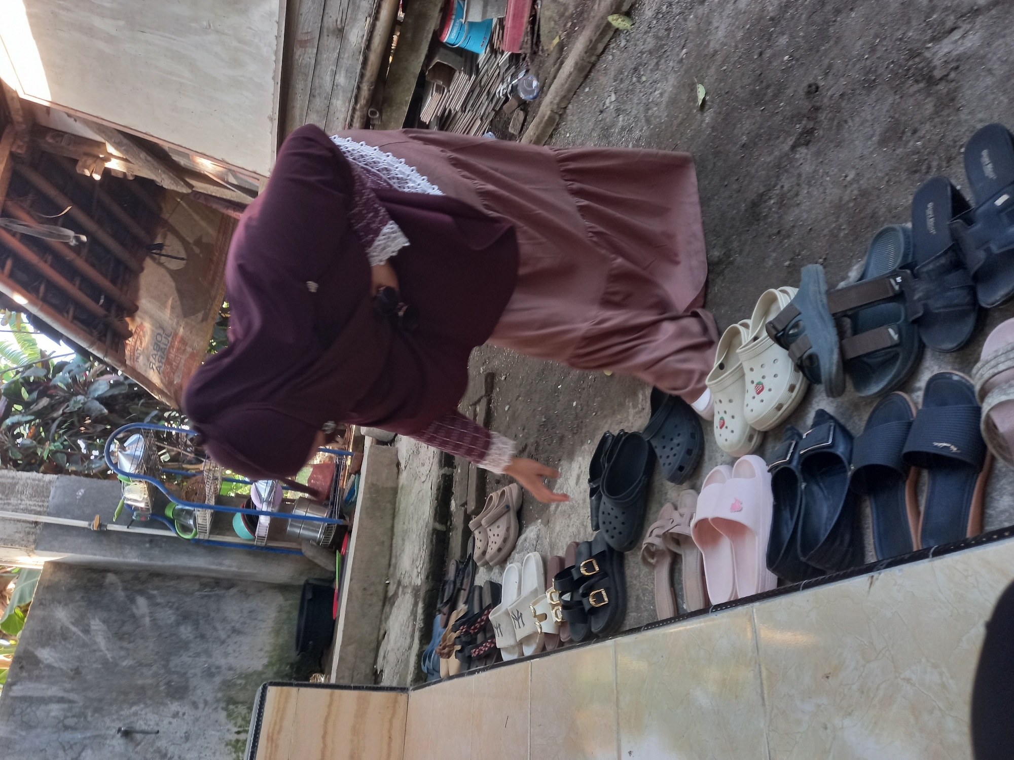In Tempos, Indonesia, a woman wearing a maroon hijab and a long, tiered brown skirt is reaching for a pair of light pink slides. She is standing in an outdoor area with a concrete floor and a partial, open-air structure with a wooden roof above. A row of various footwear, including sandals, slides, and clogs, are neatly arranged on the edge of a raised concrete ledge to her left. Behind her, a blue metal shelf unit holds several kitchen utensils, including pots and bowls. The environment appears to be a residential or semi-outdoor workspace, possibly a backyard or an area adjacent to a home.
