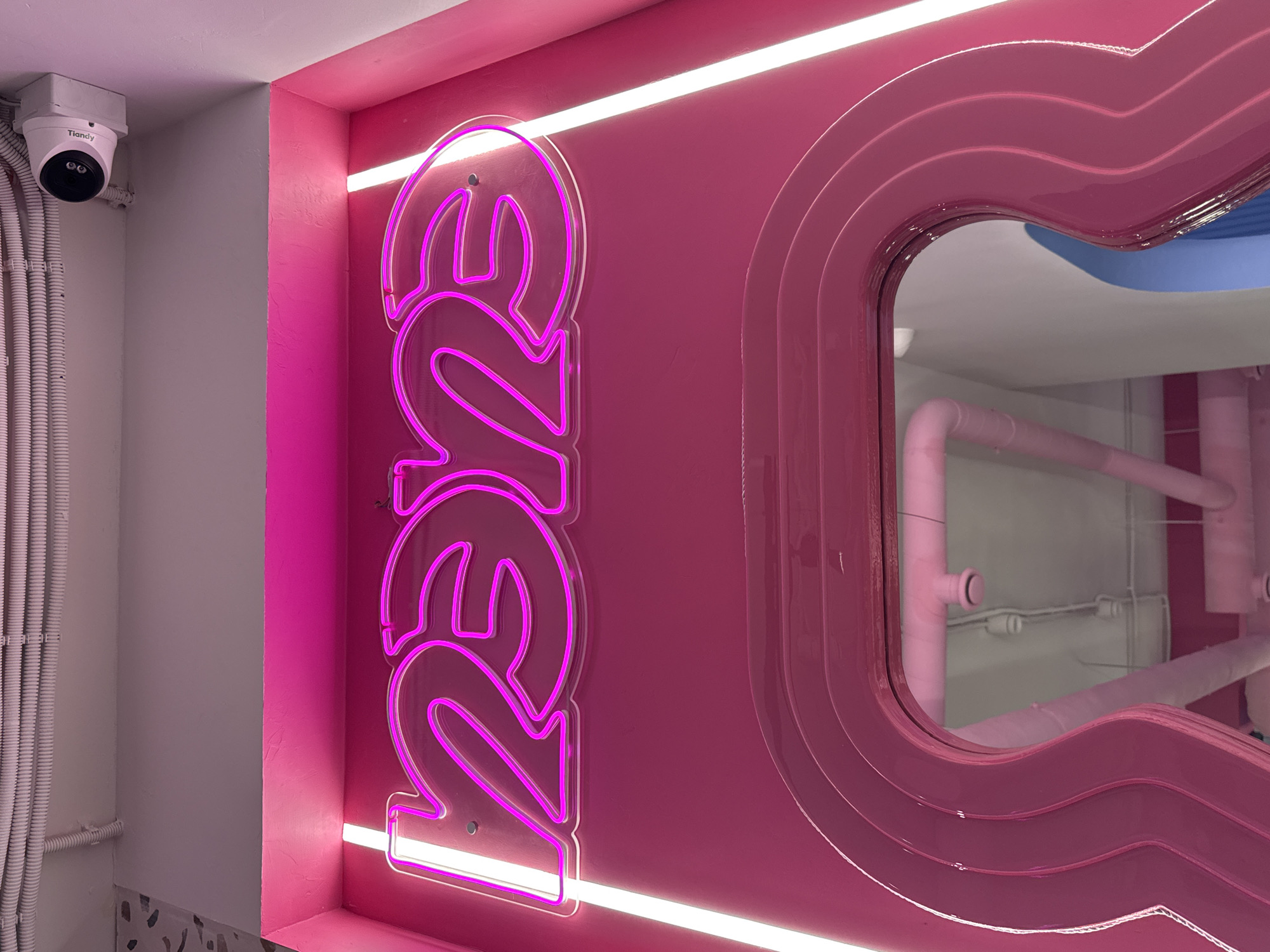 A close-up indoor shot features a pink-themed room with a neon sign, a decorative mirror, and surveillance equipment. The lower left corner shows a white wall with numerous white conduits running vertically along its surface, leading up to a white dome-shaped security camera with the brand "Tiandy" printed on it, mounted on the upper part of the wall.
To the right of the camera is a bright pink wall with a vertical neon sign displaying the text "REZE" in a stacked format with stylized, curving letters. The sign is outlined in neon pink and is mounted on a pink wall.
To the right of the neon sign is a mirror with a pink frame featuring multiple contoured layers, which creates a three-dimensional, wavy effect. The mirror reflects the interior of a room with pink pipes and a light blue ceiling detail.
The environment appears to be part of a commercial establishment or an interior design space with an emphasis on a modern aesthetic.
There is no definitive indication of this being in Sochi, Russia.