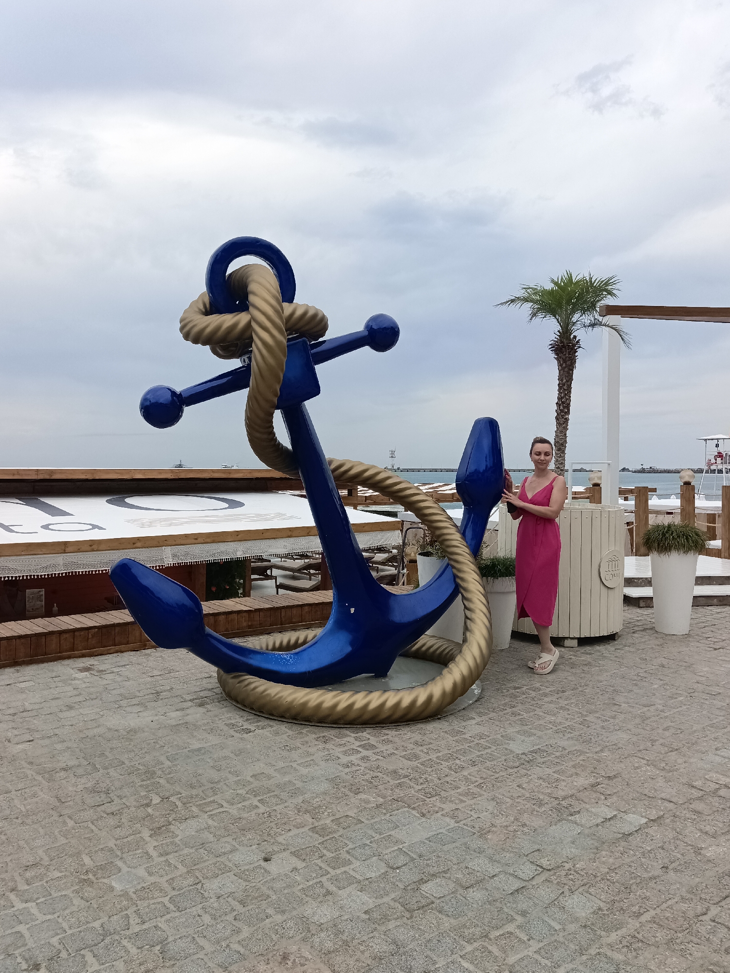 A full-shot image captures a woman standing next to a large, blue and gold anchor sculpture on a paved area near a beach. The woman, positioned to the right of the anchor, is wearing a bright pink dress and white sandals. She is holding a dark object, possibly a phone, in her right hand. The anchor sculpture is the dominant object in the foreground, with its intricate rope details visible. In the background, there are beachside structures, including wooden platforms, white awnings with a large circular logo, lounge chairs, and white planters with greenery. A palm tree stands tall behind the woman. The sea and a cloudy sky form the backdrop, with a distant view of structures on the water and a lifeguard tower on the beach. The scene appears to be set in Sochi, Russia, likely at a beach resort or promenade.