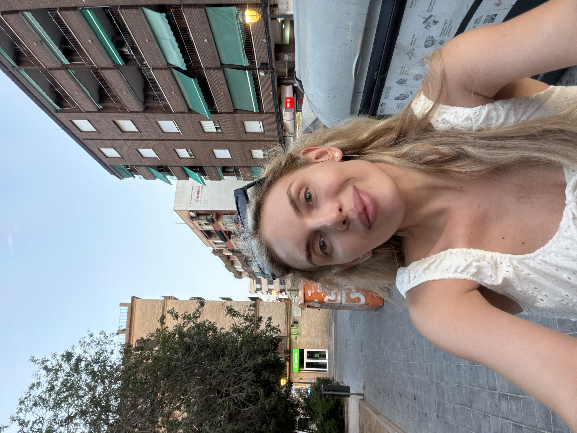 A young woman with blonde hair is taking a selfie in an urban environment. She is wearing a white, eyelet-detailed top. Her expression is a slight smile. She is positioned in the foreground, with a street and buildings in the background. To her left, there is a tree, a lamppost, a green awning over a shop with a sign that appears to say "Iberdrola", and a grey trash bin. To her right, there is a large, grey dumpster with some text and images visible on its side. Further in the background, there are multi-story brick buildings with balconies and green awnings. The sky above is a clear, pale blue, suggesting it is either early morning or late afternoon. The scene appears to be in the Benimàmet-beniferri district of Valencia, Spain, given the presence of the "Iberdrola" sign, a Spanish energy company.