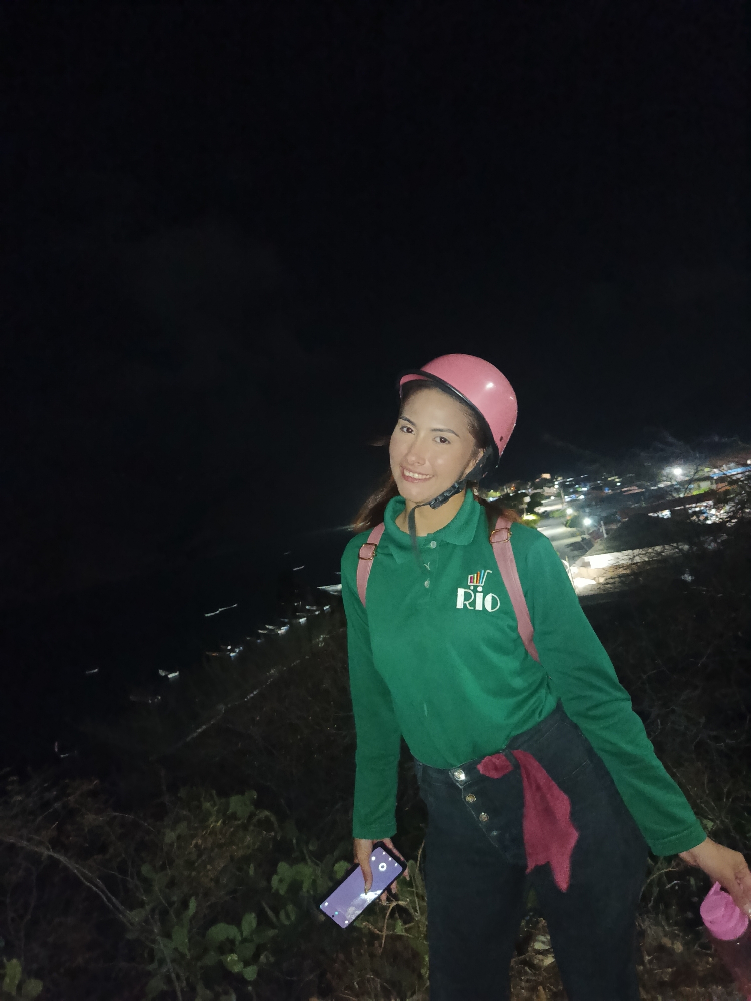 A woman is standing on a hill overlooking the city of Juan Griego, Venezuela at night. She is wearing a pink helmet, a green polo shirt with a "Rio" logo, black pants, and a pink backpack. She is holding a cell phone in her left hand and a pink water bottle in her right hand. In the background, the city lights of Juan Griego are visible, and there is dark sky above.