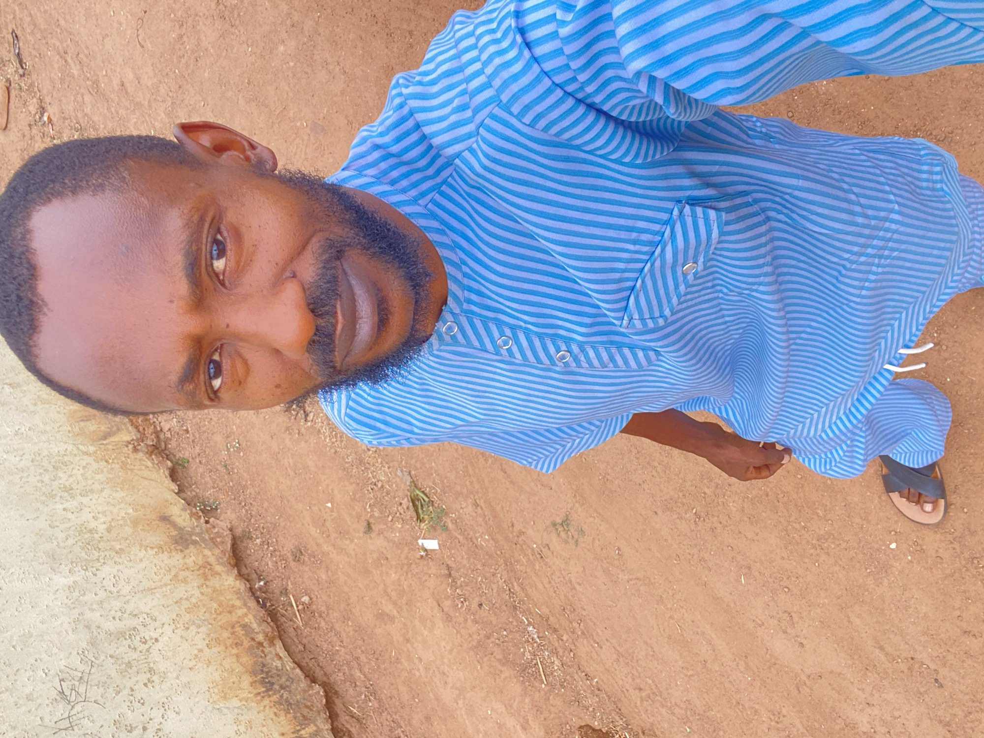 A man stands on a dirt surface in Sabon Tasha, Nigeria. He wears a light blue striped shirt with buttons and a pocket. He is looking at the camera. A sandal is visible on his foot. A concrete surface is visible to the left of the man.