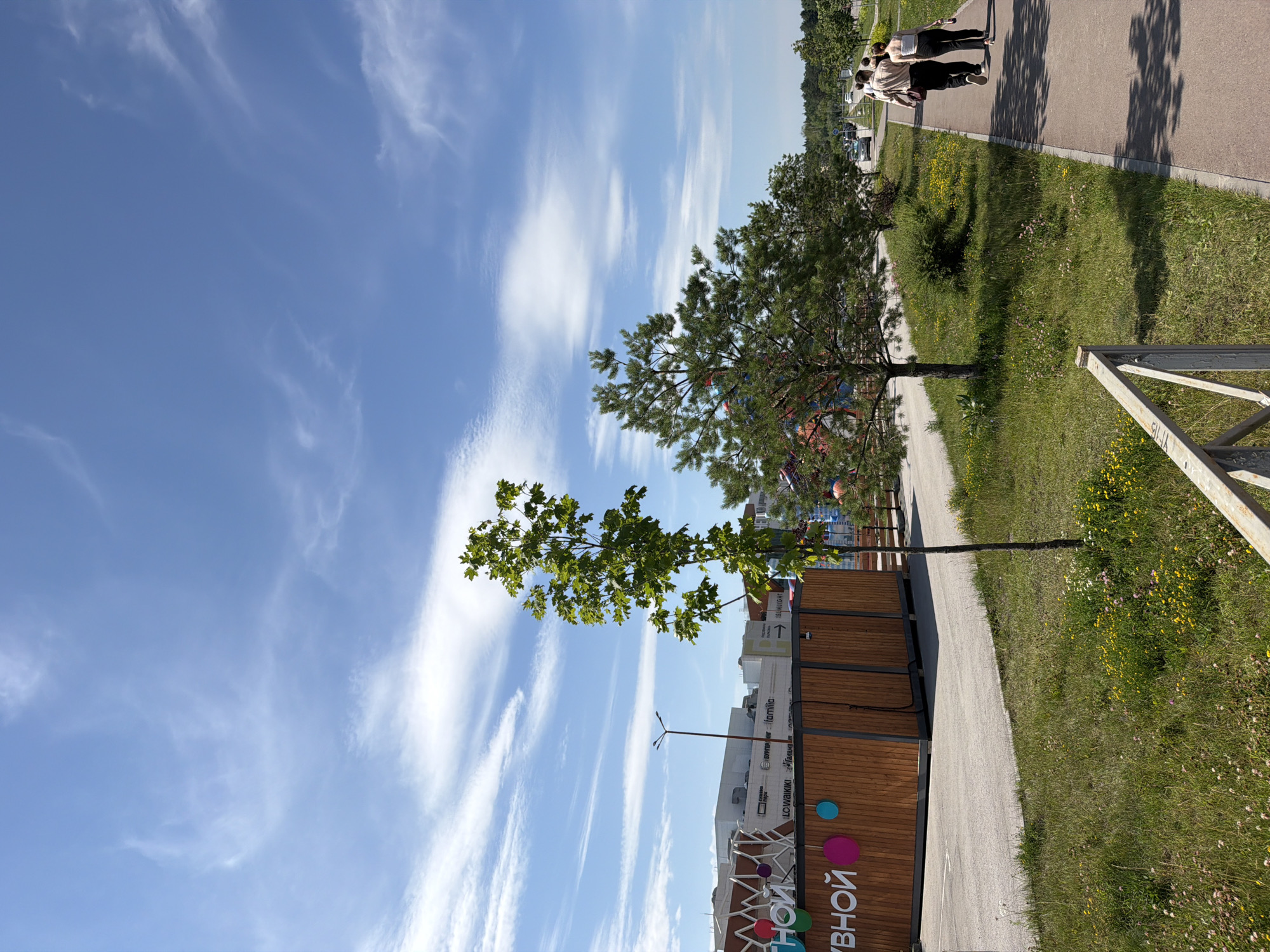 The image shows a wide outdoor pathway bordering a grassy area with wildflowers. Two individuals, a man and a woman, are walking away from the camera on the pathway. In the background, a modern building complex with a wooden facade and signage is visible, along with a street with cars and trees lining the horizon. The sky above is blue with scattered white clouds. This scene is located at the "Gorodskoy Sad" park in Perm, Russia, specifically near the park's entrance.
