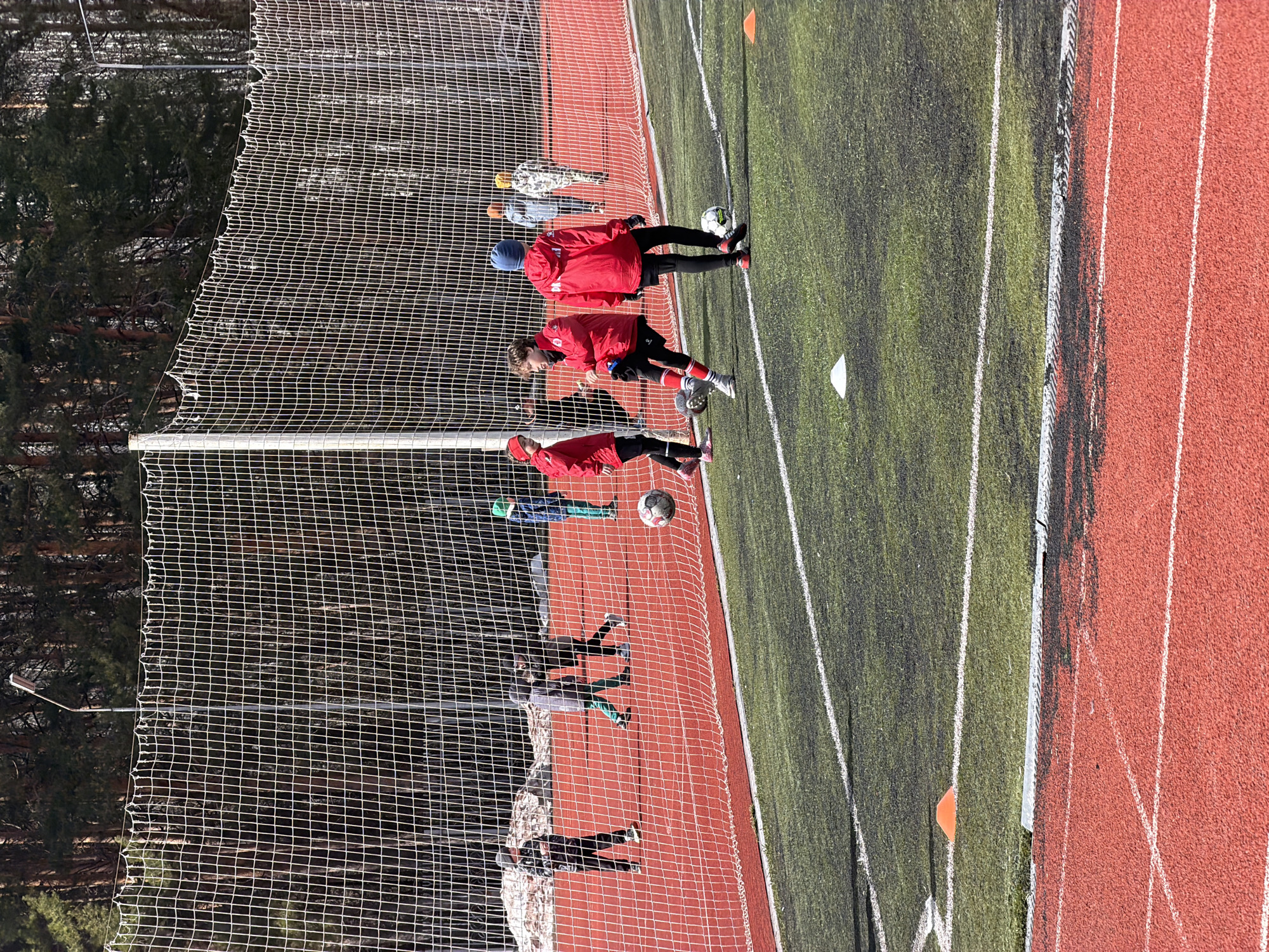 The image shows a group of young people in red jackets practicing soccer on a field in what appears to be the city of Chaikovsky, Russia, given the presence of pine trees typical of the region. The field consists of artificial green turf next to a red running track. A net stands on the side, presumably to prevent soccer balls from exiting the field. Several players can be seen interacting with soccer balls.