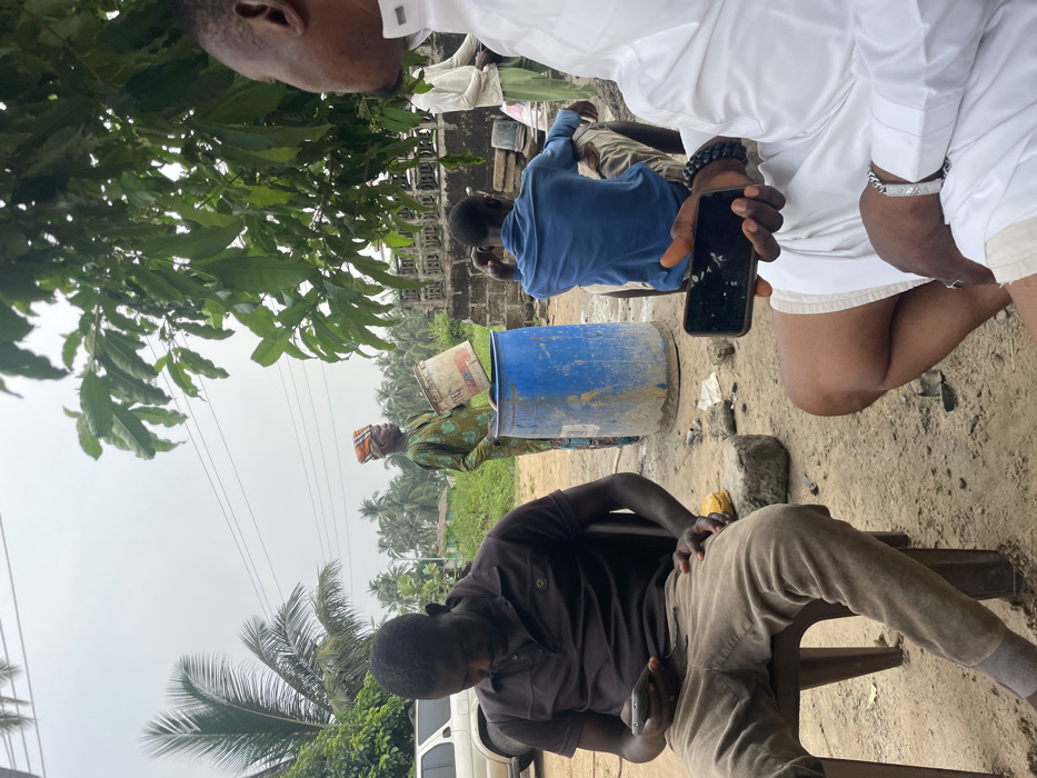 The image depicts an outdoor scene in Ganyingbo, Nigeria, under an overcast sky. The foreground shows an unpaved, light brown dirt ground with scattered debris.
Five adult males are present. In the immediate foreground left, a dark-skinned man wearing a dark brown polo shirt and light brown pants sits on a brown plastic chair, looking down at a dark-colored smartphone held in both hands. To his right, a partial view of another dark-skinned man shows him seated, wearing a white robe-like garment and white shorts. He holds a black smartphone with a cracked screen in his left hand and wears a black beaded bracelet on his left wrist and a silver-colored chain bracelet on his right wrist.
In the midground, a dark-skinned man in a patterned green long-sleeved garment and an orange and brown headwrap stands, facing right. He holds a white plastic bucket tilted over an upright blue plastic barrel, appearing to pour contents into it. Behind the barrel, another dark-skinned man in a blue long-sleeved shirt and light-colored pants is seated on a low object, facing away from the viewer. Further in the background right, partially obscured by green foliage and a concrete block wall, a fifth dark-skinned man in a light-colored long-sleeved shirt stands.
The background features lush green vegetation, including numerous palm trees and other broad-leafed trees. A white SUV is partially visible in the far left background. Overhead power lines traverse the grey, cloudy sky.