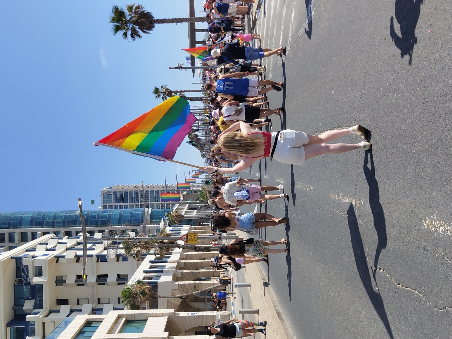 The image captures a sunny outdoor scene in Tel Aviv, Israel, likely during a Pride event, given the prominent display of a rainbow flag. A young woman in the foreground, facing away from the camera, is holding the rainbow flag high with both hands, her arms raised. She is wearing a red halter top and white shorts. The flag, with its vibrant stripes of red, orange, yellow, green, blue, and purple, is a focal point.
Behind her, a crowd of people is walking down a paved street. Most are facing away from the camera, suggesting they are moving in the same direction as the woman with the flag. The people are dressed in casual summer attire, consistent with a warm climate. Some are carrying smaller flags or wearing festive accessories.
The environment shows an urban setting with modern, multi-story buildings lining the left side of the street. These buildings have a contemporary architectural style with large windows and balconies. Palm trees are visible interspersed between the buildings and along the street, indicating a coastal or Mediterranean city. The sky is clear and blue. The street itself is asphalt with visible white lines marking lanes. Shadows of people and possibly structures are cast on the road surface, indicating the time of day is likely midday. In the distance, more buildings and palm trees are visible, and the street appears to be a wide boulevard.