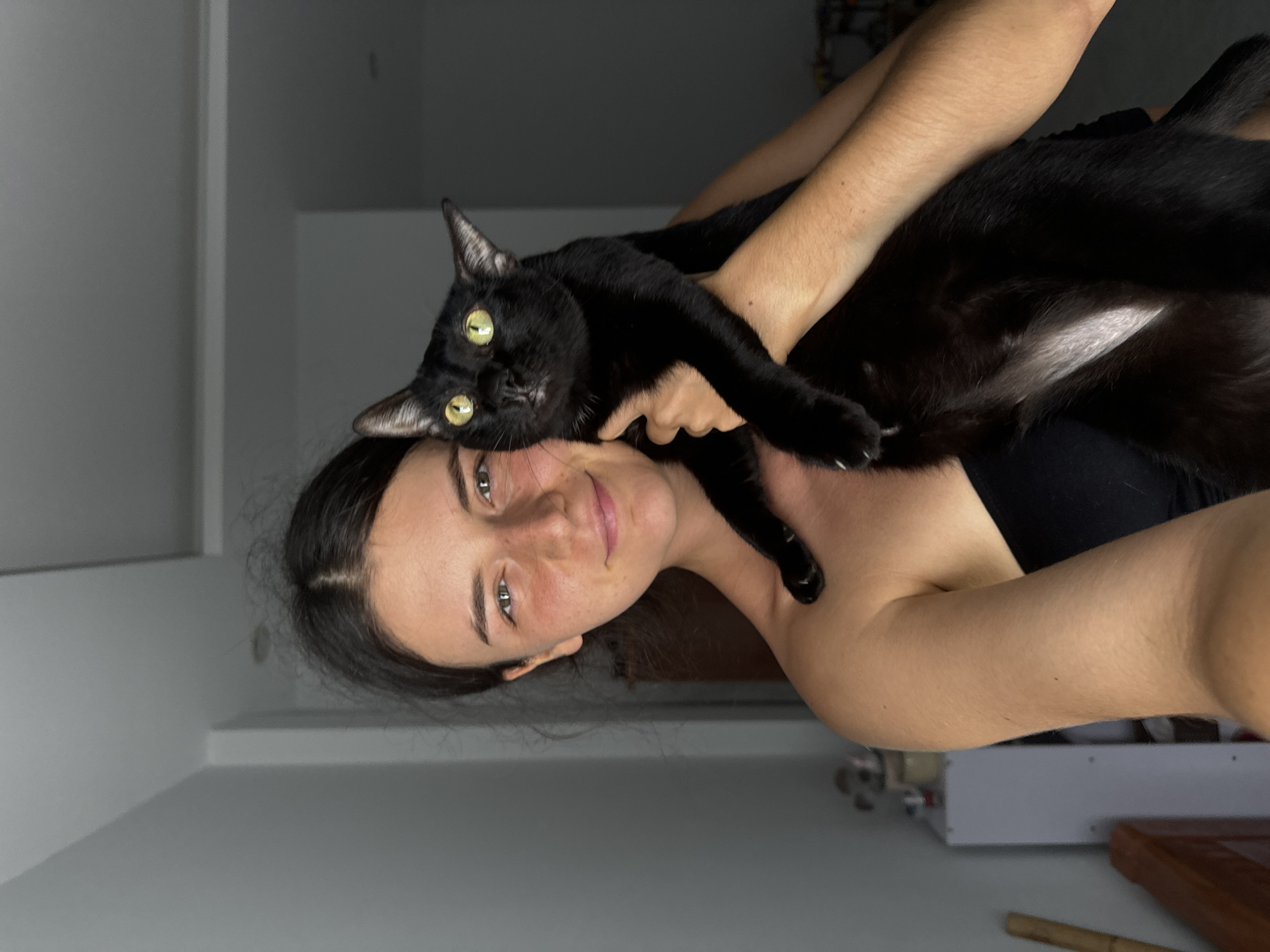A young woman with dark hair and fair skin is holding a black cat. She is wearing a black strapless top. The cat has bright green eyes and is looking directly at the camera. They are indoors, in what appears to be a room with white walls. A white cabinet and some wooden furniture are visible in the background. The lighting suggests it is either evening or the room is dimly lit. The image is a portrait of the woman and her cat. There is no indication of the location being in Mũi Né, Vietnam.