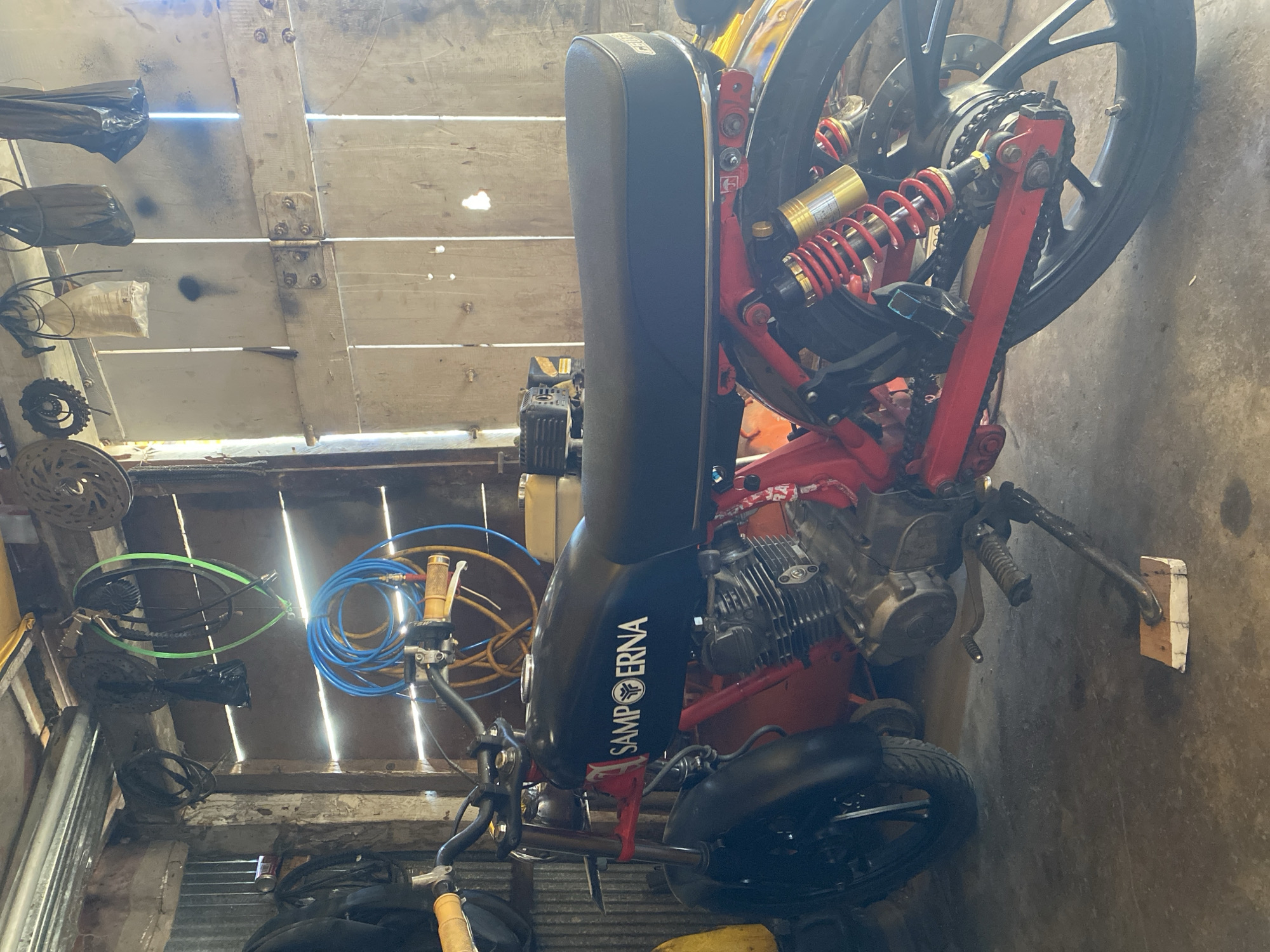 The image depicts a custom motorcycle parked inside a rustic shed, likely located in Bantaeng, Indonesia. The motorcycle is predominantly black with red accents, featuring a visible "SAMPOERNA" logo on its fuel tank. Red suspension components and a red frame are prominent. The shed's interior is characterized by wooden walls and a corrugated metal side. Various items are hung from the ceiling, including hoses, cables, and bags. The floor is concrete. The lighting suggests it is daytime, with sunlight filtering through gaps in the wooden structure. No people are visible in the image.
