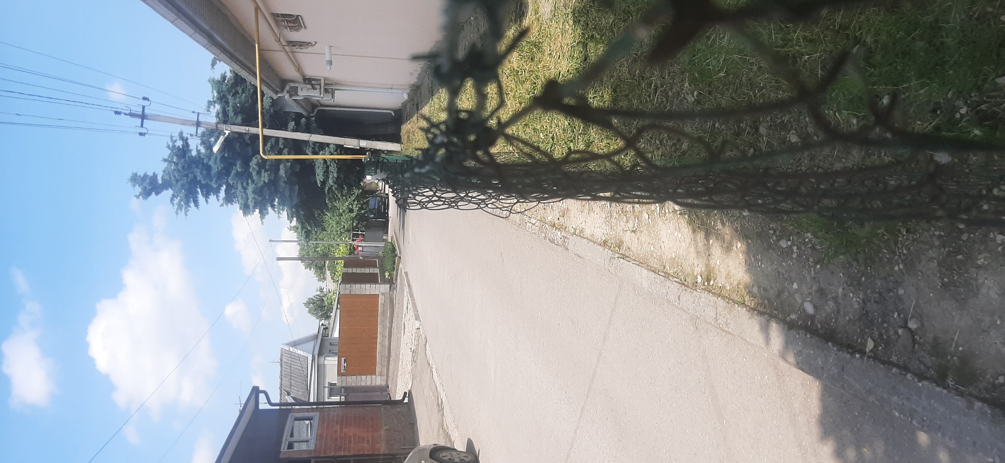 The image depicts a street scene in Yessentukskaya, Russia, on a sunny day. A narrow asphalt road runs through a residential area, bordered by houses and fences. A prominent barbed wire fence runs along the right side of the road in the foreground, partially obscuring the view of the adjacent grassy area and a light-colored building. In the background, a dark-colored car is parked on the side of the road. Overhead, power lines and a yellow gas pipe are visible against a blue sky with scattered white clouds. A tall evergreen tree stands near a utility pole. The overall impression is of a quiet, suburban street.