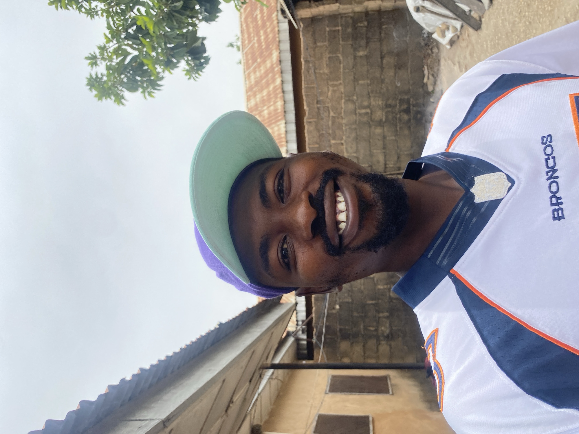 A man with dark skin, a beard, and a mustache is captured in a cheerful outdoor portrait, smiling broadly with his teeth showing, and looking directly at the camera. He is wearing a two-toned baseball cap with a light green brim and a purple crown. His attire includes a white sports jersey with blue and orange accents, and the word "BRONCOS" is clearly visible in blue text with an orange outline on the right side of his chest. A small, patterned emblem is also present on the upper left chest area of the jersey.
The image is taken from a low angle, with the man's head and shoulders filling much of the frame. The background reveals an overcast sky, suggesting a cloudy day. Above him, green foliage from a tree is visible. Architectural elements behind him include corrugated metal roofing, indicative of local construction, and walls made of what appears to be concrete blocks or bricks, suggesting buildings in a residential or commercial area. Further back, another structure with dark rectangular features, possibly windows, is discernible. The scene implies a casual setting in Barakin Akawo, Nigeria, with the man seemingly posing for a selfie or a close-up shot.
