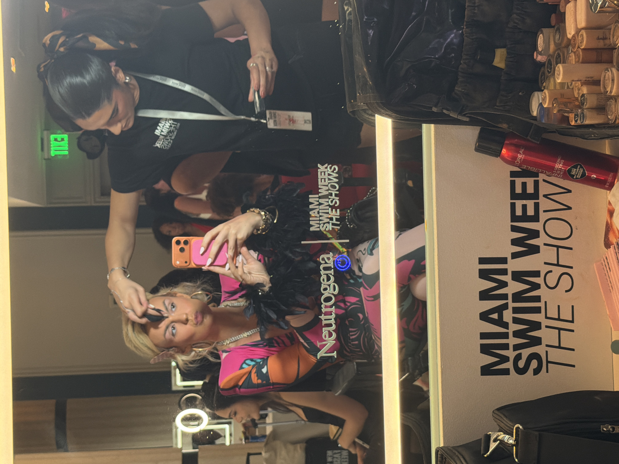 Here is a description of the image:
This photo depicts a scene from Miami Swim Week in Miami Beach, Florida, USA. A woman with blonde hair, gold hoop earrings, and a colorful dress with a feather boa takes a selfie in a mirror while getting her makeup done. The makeup artist, who has dark hair tied in a bun, is applying makeup with a brush. The reflection in the mirror shows the woman's phone, adorned with a Neutrogena logo. In the background, makeup supplies are visible, and a sign reads "MIAMI SWIM WEEK THE SHOWS." The photo conveys the behind-the-scenes preparations at a fashion event in Miami Beach.
