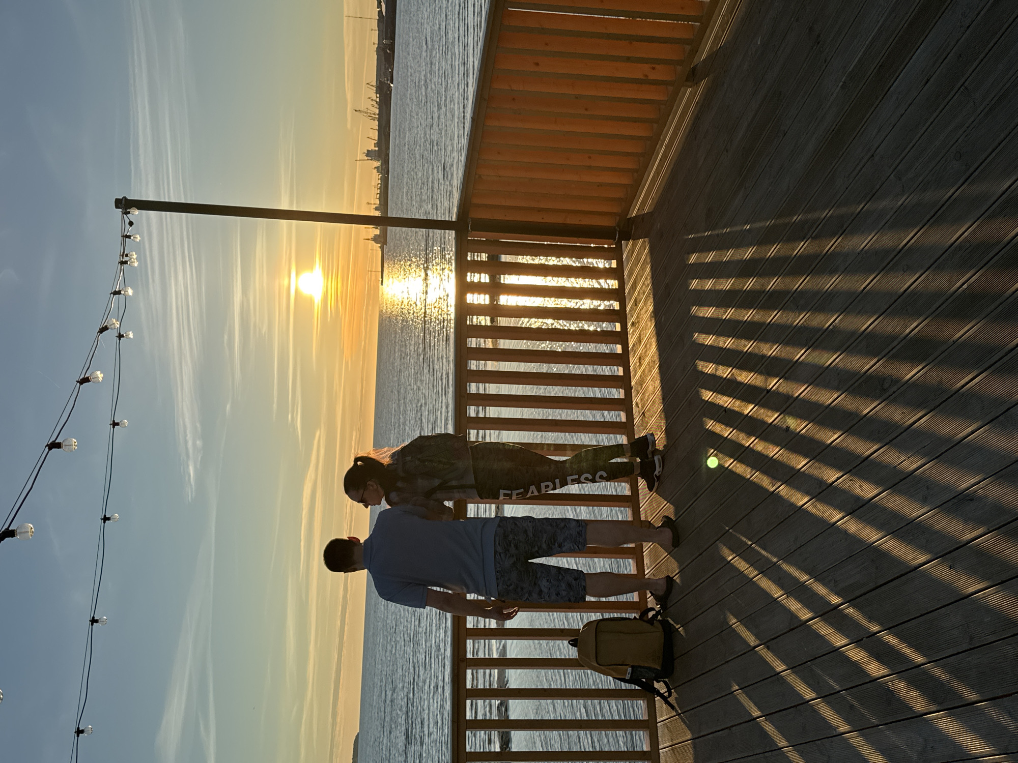 The image depicts a couple standing on a wooden deck overlooking the sea during sunset. The sun is low on the horizon, casting a warm glow and reflecting on the water. The couple is viewed from behind, with the male figure wearing a blue polo shirt and patterned shorts, and the female figure wearing a patterned top and leggings, and a backpack. A yellow backpack rests on the deck near the man's feet. A string of lights hangs overhead, and a wooden railing separates the deck from the water. In the distance, a port with cranes and ships is visible. The deck is made of wooden planks, and the setting sun creates long, striped shadows. The scene is likely located at a waterfront area in Tallinn, Estonia, possibly near the Tallinn Port or a promenade with a view of the harbor.