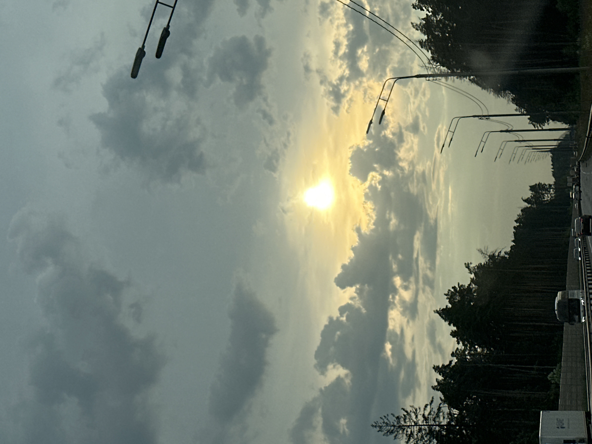 This is a photograph taken from a vehicle on a highway, likely in or near Khliupino, Russia. The scene depicts a cloudy sky with the sun peeking through, casting a bright glow. A dense forest of pine trees lines the right side of the highway, with a few trees visible on the left. Several streetlights are positioned along the road, and power lines are strung between them. The highway is a multi-lane road with traffic moving in the same direction. Several vehicles, including trucks and cars, are visible on the road. The overall atmosphere is somewhat overcast, with the sun providing a dramatic contrast against the dark clouds and trees.