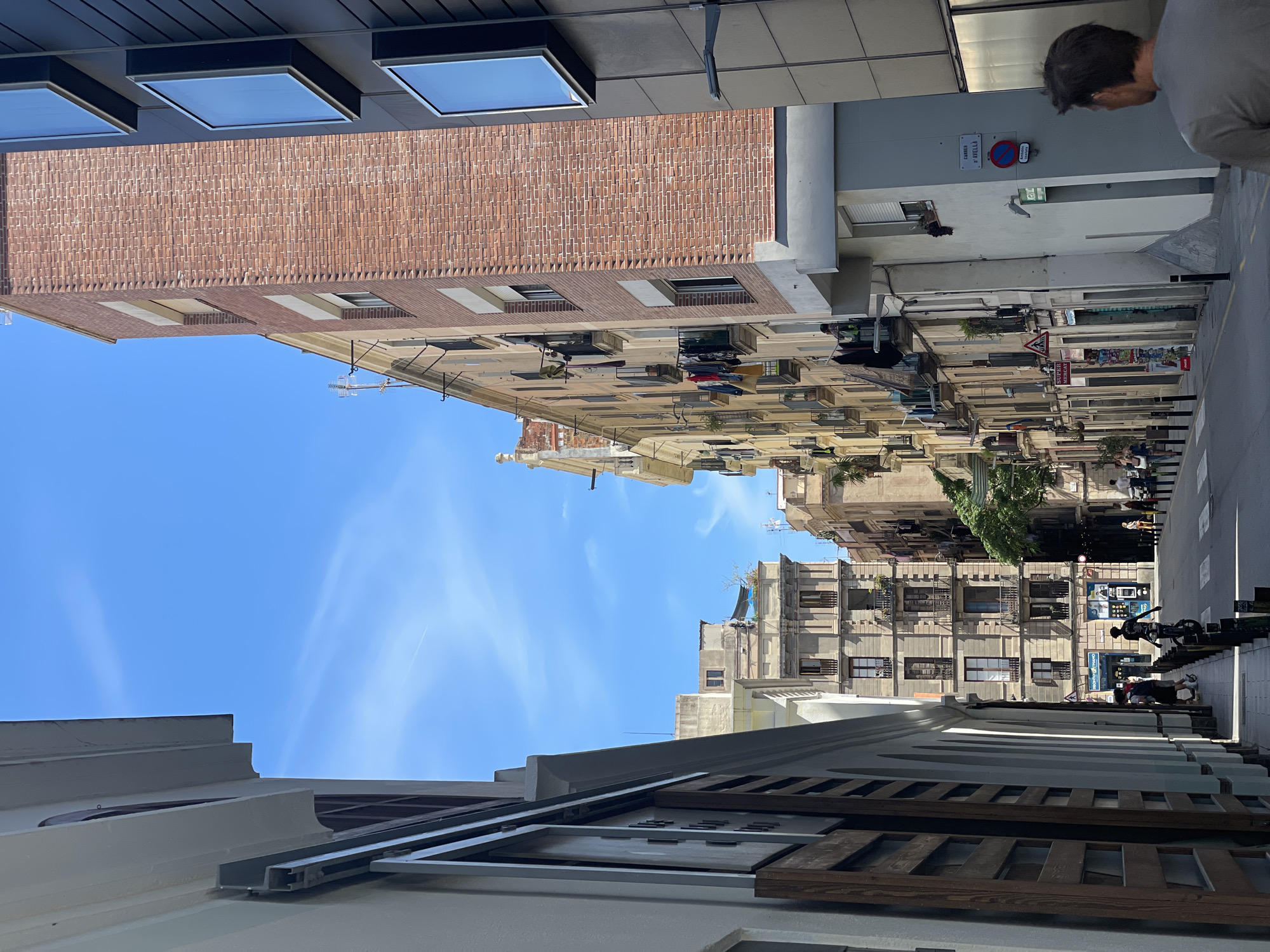 The image displays a narrow urban street, identified by a sign as Carrer d'Avellà, located in Barcelona, Spain. The scene is viewed looking down the street on a bright, sunny day with a clear blue sky featuring minimal, wispy clouds and a distant aircraft contrail.
On the immediate left, a light-colored building facade with dark-framed openings and a pronounced decorative cornice extends into the foreground. Further down the left side, a row of multi-story buildings, predominantly residential, features balconies with traditional shutters and some potted plants.
On the right side of the street, a tall building constructed of reddish-brown brick with horizontal bands of lighter-colored material and white-framed windows is prominent. Adjacent to it, a modern building with a dark panel facade displays multiple vertically stacked, rectangular, illuminated screens. Below this, a lighter-colored building features a street sign reading "CARRER D'AVELLÀ" and a circular no-parking sign. Laundry is visible hanging from several balconies on the buildings further down the right side.
The street is paved, with a pedestrian sidewalk running along the left, delineated by black bollards. A white crosswalk is marked in the midground. Several individuals populate the scene: a man in a grey t-shirt is seen from behind in the bottom right foreground, walking away. Two people walk on the left sidewalk, also moving away from the viewer. A person rides a unicycle in the street. Additional pedestrians are visible on the right sidewalk further down the street. Commercial signage, including "SUPER" and "ATM," is visible on storefronts at ground level. Antennas are present on rooftops, and sparse green foliage appears between distant buildings.