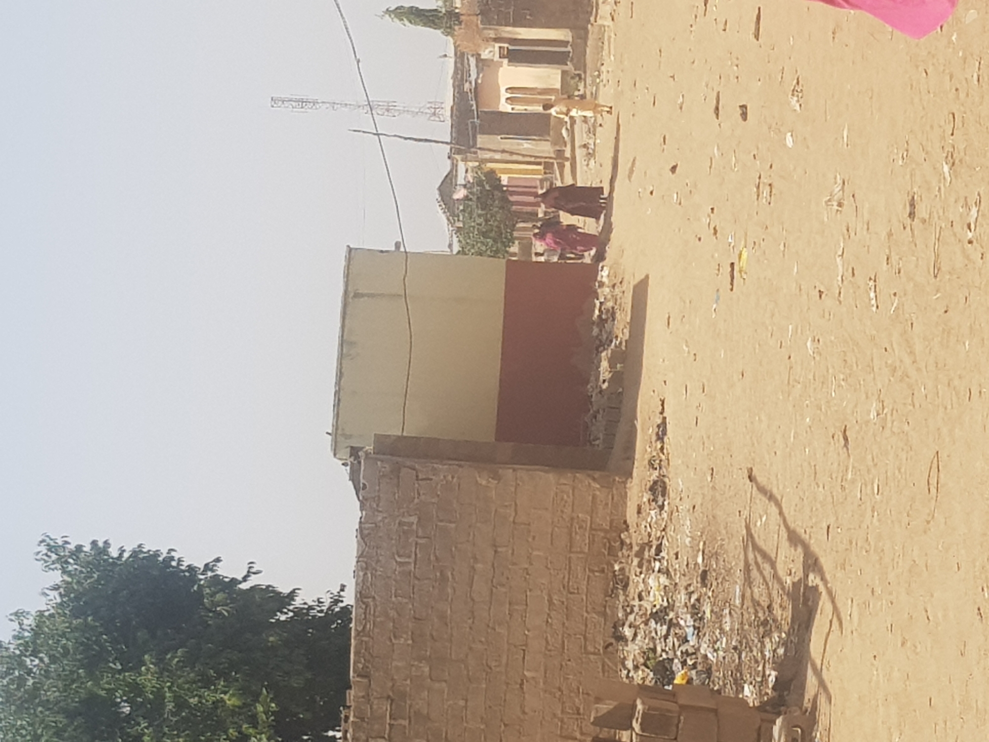 The image shows a street scene in Dabawa, Nigeria. The foreground features a brick wall and litter-strewn ground. Further down the dirt road, two women in colorful robes are walking together. Across the street, a dog is standing near a building. In the background, a communication tower rises above the buildings. The sky is bright and hazy.