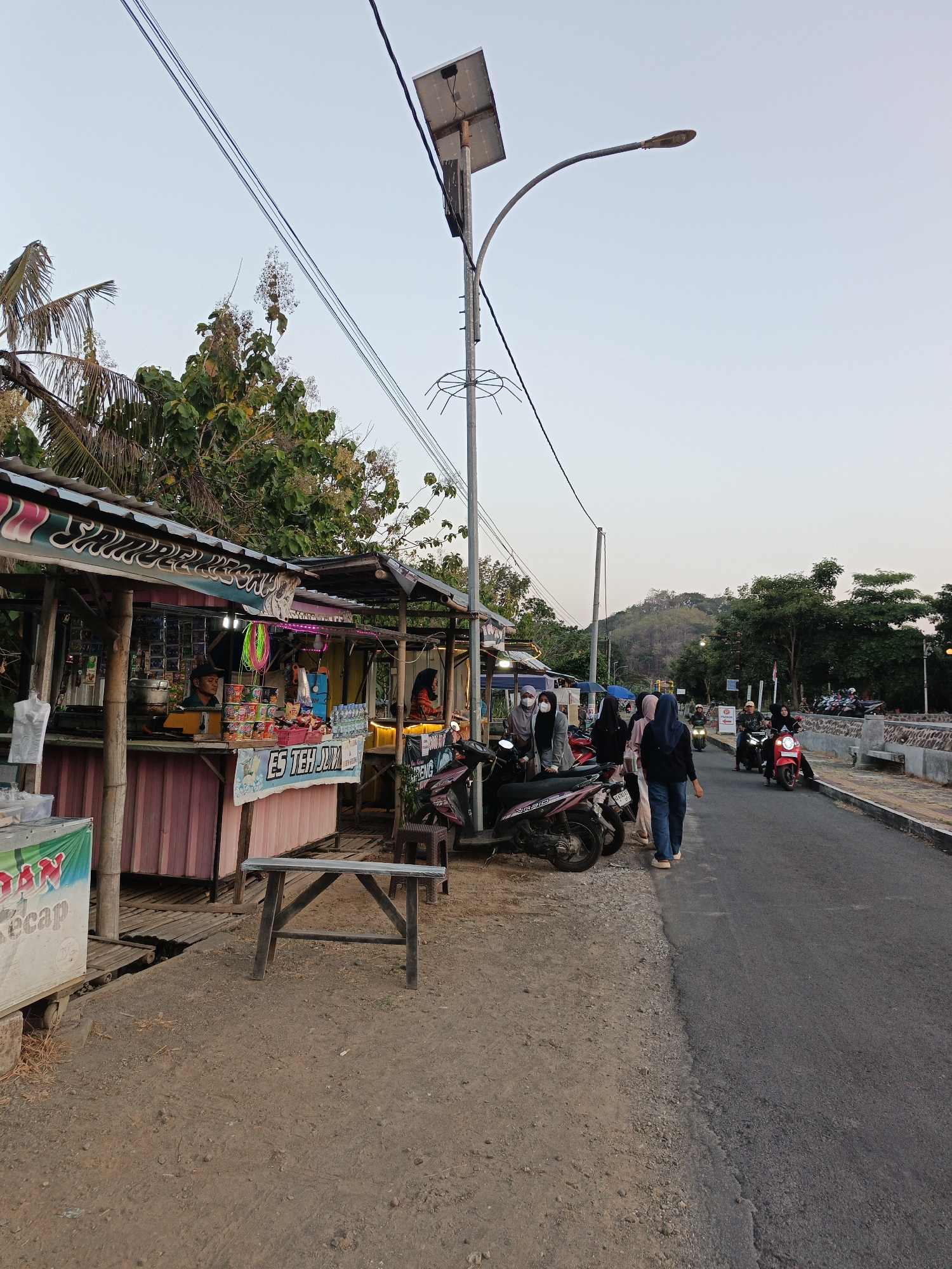 This is a medium shot, taken at dusk, of a street scene in Krakitan, Indonesia, featuring small roadside stalls and people on motorbikes. The foreground is dominated by a dirt and gravel area with a simple wooden table and some debris. To the left, a small, open-air stall with a corrugated metal roof displays various items for sale, including drinks and snacks. A vendor is visible inside the stall. Several people, mostly women wearing headscarves, are gathered near the stall and on motorbikes. One woman in a blue headscarf and jeans is walking away from the camera on the asphalt road. To the right, the road is busy with several motorbikes, some of which are ridden by men. A stone wall and more trees are visible in the background, and a hill with vegetation rises in the distance. The sky is clear and a pale orange hue, indicative of sunset. A solar-powered street lamp with a curved arm is positioned prominently in the upper left quadrant of the image.