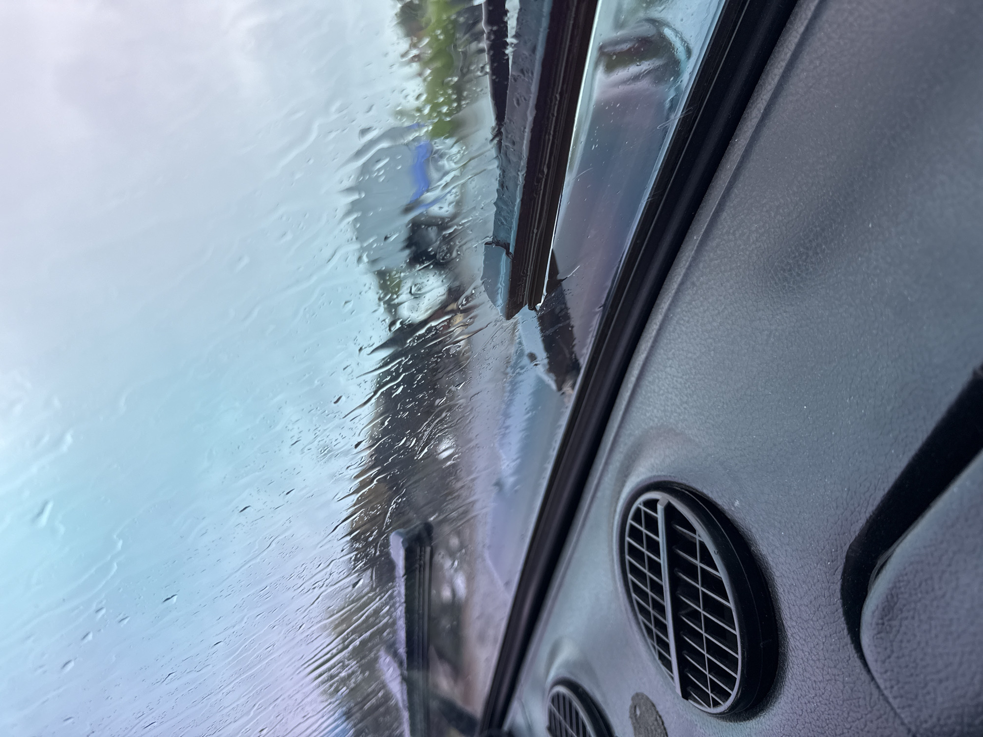 The image is taken from inside a vehicle, likely a car, looking out through a rain-streaked windshield. Windshield wipers are partially visible, with one wiper blade resting on the glass. Outside the vehicle, the scene is obscured by the rain, though vague shapes of trees or buildings can be discerned. The interior of the vehicle is visible, including part of the dashboard and circular air vents with horizontal grilles. The color of the interior is dark, possibly black or dark gray. The image provides no information about the location being Chaikovsky, Russia.