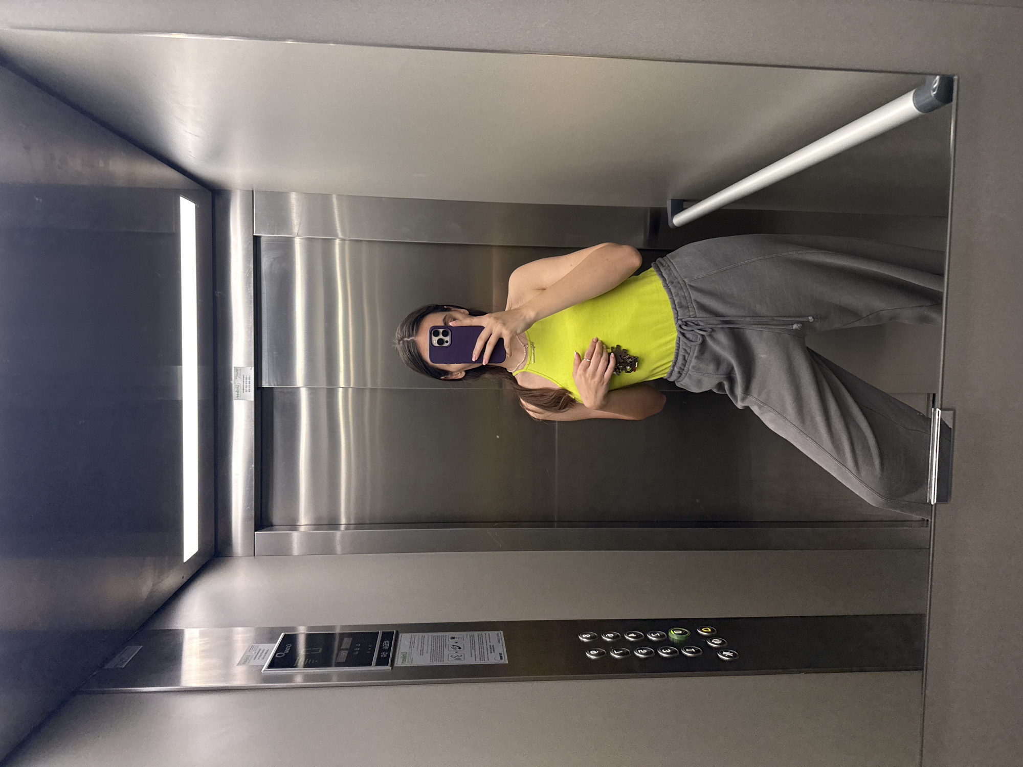 The image shows a selfie taken in an elevator. A young woman is holding a smartphone with a purple case, covering her face. She is wearing a bright yellow tank top and gray sweatpants. Her hair is dark and shoulder-length. The elevator has stainless steel walls and a horizontal grab bar. The control panel with buttons is visible, as well as the floor level indicator. No information is available in the image that could place this scene in Plovdiv, Bulgaria.