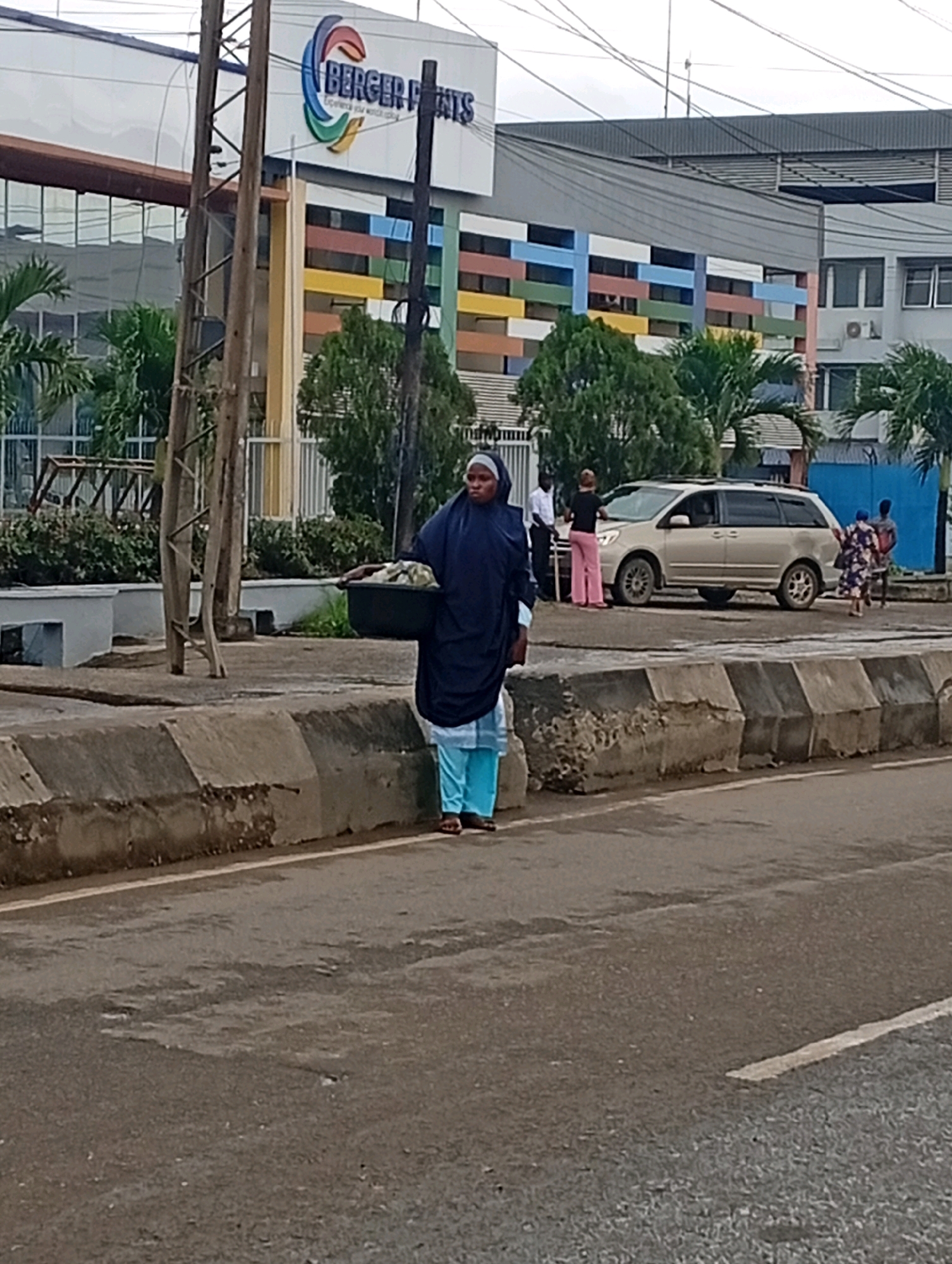 In Ikeja, Nigeria, a woman wearing a dark blue hijab and a light blue garment walks on the side of a paved road. She is carrying a dark basin filled with what appears to be laundry. Behind her, a tan minivan is parked on the side of the road. Several other people are visible in the background, including a person in a white shirt and pink pants, and two individuals near the back of the minivan. The scene is set against the backdrop of a modern building with a colorful facade, featuring horizontal bands of blue, yellow, red, and white. Utility poles and wires are prominent in the foreground, suggesting an urban environment. A concrete divider runs along the edge of the road, separating it from a landscaped area with trees and bushes. The sky appears overcast.