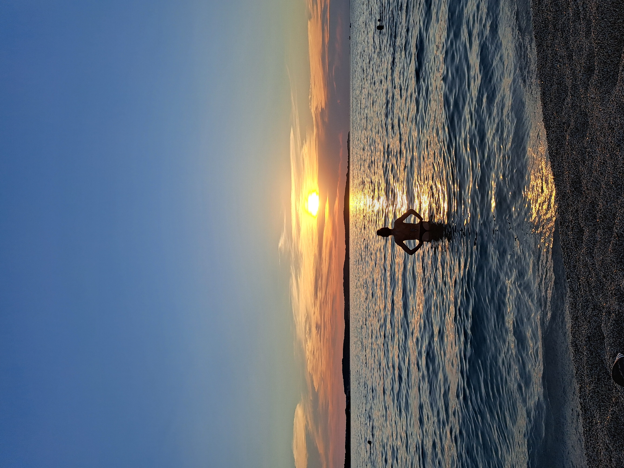 A person stands waist-deep in the ocean at sunset. The sun, a bright orange orb, is low on the horizon, casting a golden glow on the water and sky. The sky is a gradient of blue and orange, with wispy clouds tinged with pink and yellow. The water is calm with gentle ripples, reflecting the sunlight. The shore is visible on the right side of the image, covered in dark pebbles. The person is silhouetted against the setting sun, their back to the camera, with their arms bent at the elbows. The overall mood is peaceful and serene, capturing a moment of quiet enjoyment of nature's beauty. The time of day is clearly sunset, and the weather appears to be clear with some cloud formations. There are no visible objects or text in the image.