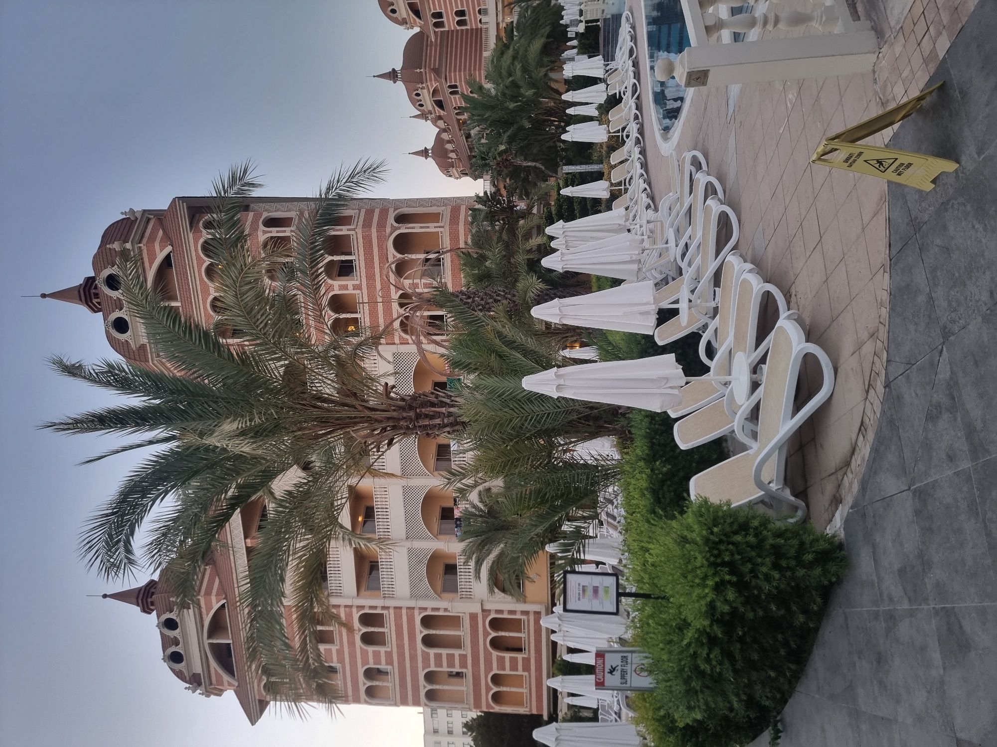 The image shows an exterior view of a hotel resort in Manavgat, Turkey, likely during the early morning or late evening, given the soft lighting. In the foreground, a row of white lounge chairs and folded umbrellas are neatly arranged on a tiled patio next to a swimming pool. A yellow caution sign indicating a "SLIPPERY FLOOR" is prominently placed on the ground. Lush green bushes border the patio.
Behind the poolside area, a tall, multi-story hotel building with ornate architecture is visible. The building features a combination of red brick and pale-colored decorative elements, with arched windows and balconies. Palm trees and other tropical foliage are interspersed around the building and the resort grounds. In the background, other sections of the hotel with domed rooftops can be seen. There are no people visible in the image. The overall scene conveys a sense of tranquility and resort ambiance.