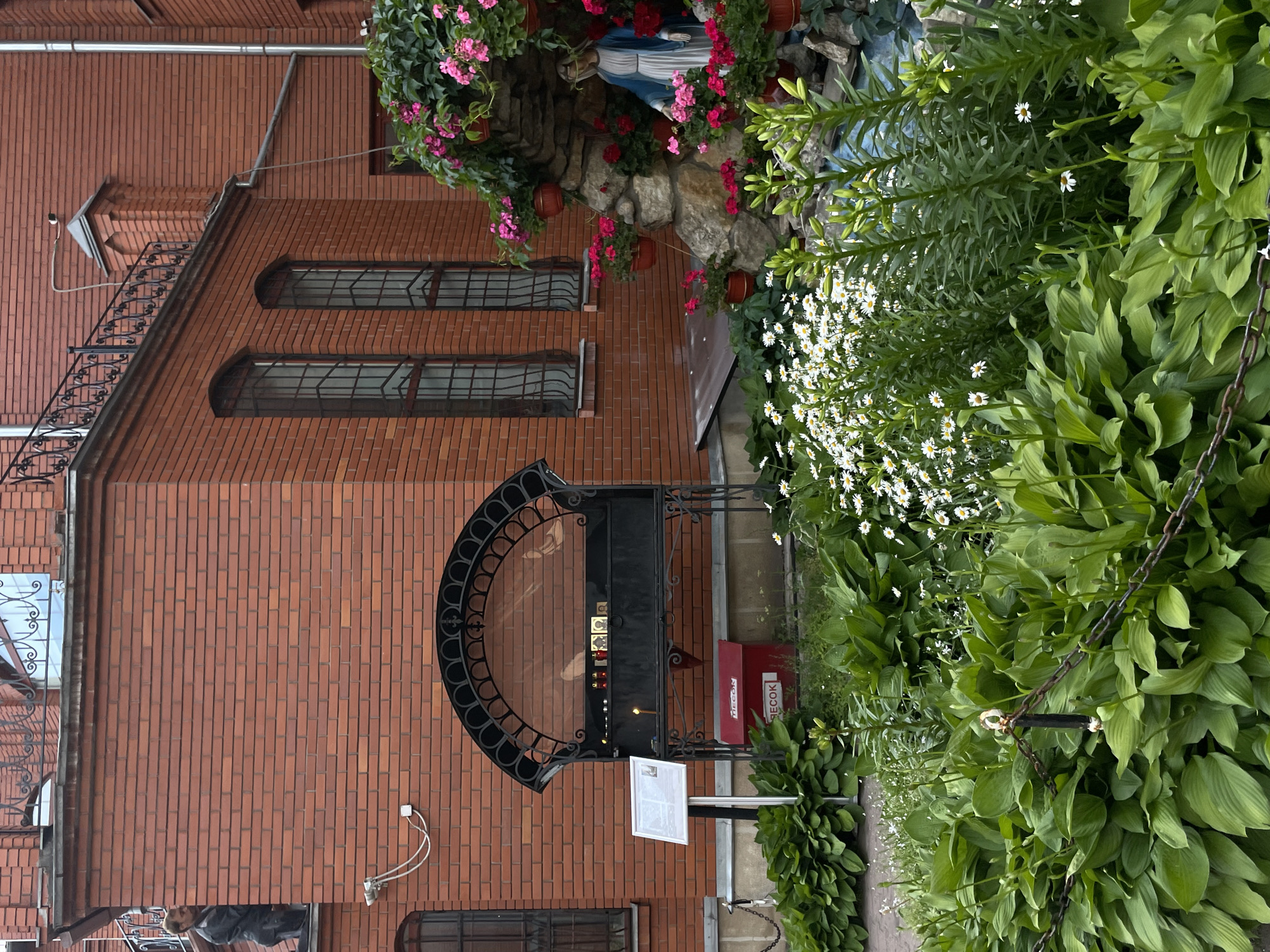 The image depicts an exterior courtyard of a brick building, possibly a religious institution, in Moscow, Russia. The building features arched windows with ornate iron grates and a decorative railing on an upper level. In the foreground, lush green hosta plants and a bed of white daisies are visible. A chain-link fence runs across the lower portion of the image, separating the garden from the viewer.
To the right, a grotto-like structure is adorned with pink and red flowers in hanging pots. A statue of the Virgin Mary, draped in a blue mantle, stands within the grotto. Adjacent to the building's wall, a black metal structure with a decorative arched top contains several lit candles and icons, suggesting a place for prayer or offerings. Below this structure, a red metal box with Cyrillic lettering, likely a donation box, is placed on the ground. To the left of the candle stand, a white information board is mounted on a stand.
In the upper left quadrant of the image, a man in a dark jacket is visible standing on a ledge near the building's edge, looking down. A security camera is mounted on the brick wall below him. The overall scene is one of a tranquil courtyard, blending architectural elements with natural beauty and religious iconography.