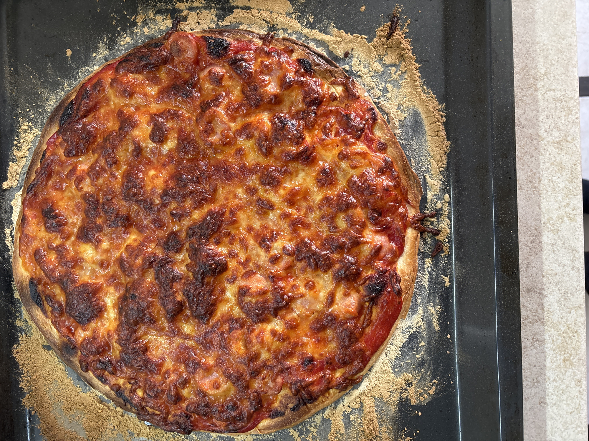 A close-up overhead shot shows a golden-brown, round pizza with melted cheese, placed on a dark baking sheet. Some of the cheese has caramelized to a darker brown, giving it a slightly charred appearance. The pizza crust is visible around the edges and is also golden brown. A scattering of fine, light brown crumbs is visible on the baking sheet, both under and around the pizza. The baking sheet itself is dark and appears to be made of metal. The bottom portion of the image shows a countertop with a speckled pattern of beige and white. The scene is well-lit, suggesting it might be indoors. There are no people or animals present in the image. The image does not contain any elements that would allow for the determination of its location in Bila Tserkva, Ukraine.