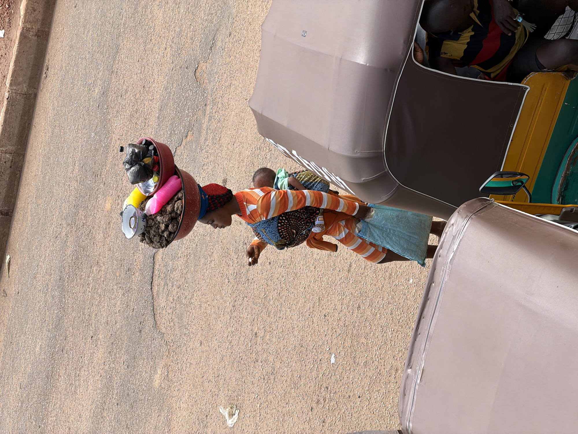In Bukuru, Nigeria, a woman is depicted from a high angle, carrying a basket filled with various items. She is dressed in a vibrant orange and white striped garment, with a patterned fabric tied around her waist. Her head is adorned with a red headscarf. On her shoulder, she carries a dark red bowl overflowing with what appears to be earth or natural material, and some colorful objects, including a pink cylindrical item and a yellow item. Her face is not clearly visible, but her posture suggests she is in motion, possibly walking.
Behind her, a portion of a vehicle is visible, with a beige canopy and hints of yellow and green paint. Inside the vehicle, another person can be seen, wearing a dark shirt with horizontal stripes of yellow, red, and black. The setting appears to be an outdoor, paved area, likely a street or marketplace, with a textured, sandy ground. The lighting suggests it is daytime, possibly sunny, casting shadows that are not prominently visible. The overall scene captures a moment of daily life and commerce, highlighting the woman's burden and her engagement in an unspecified activity. No text is clearly legible in the image.