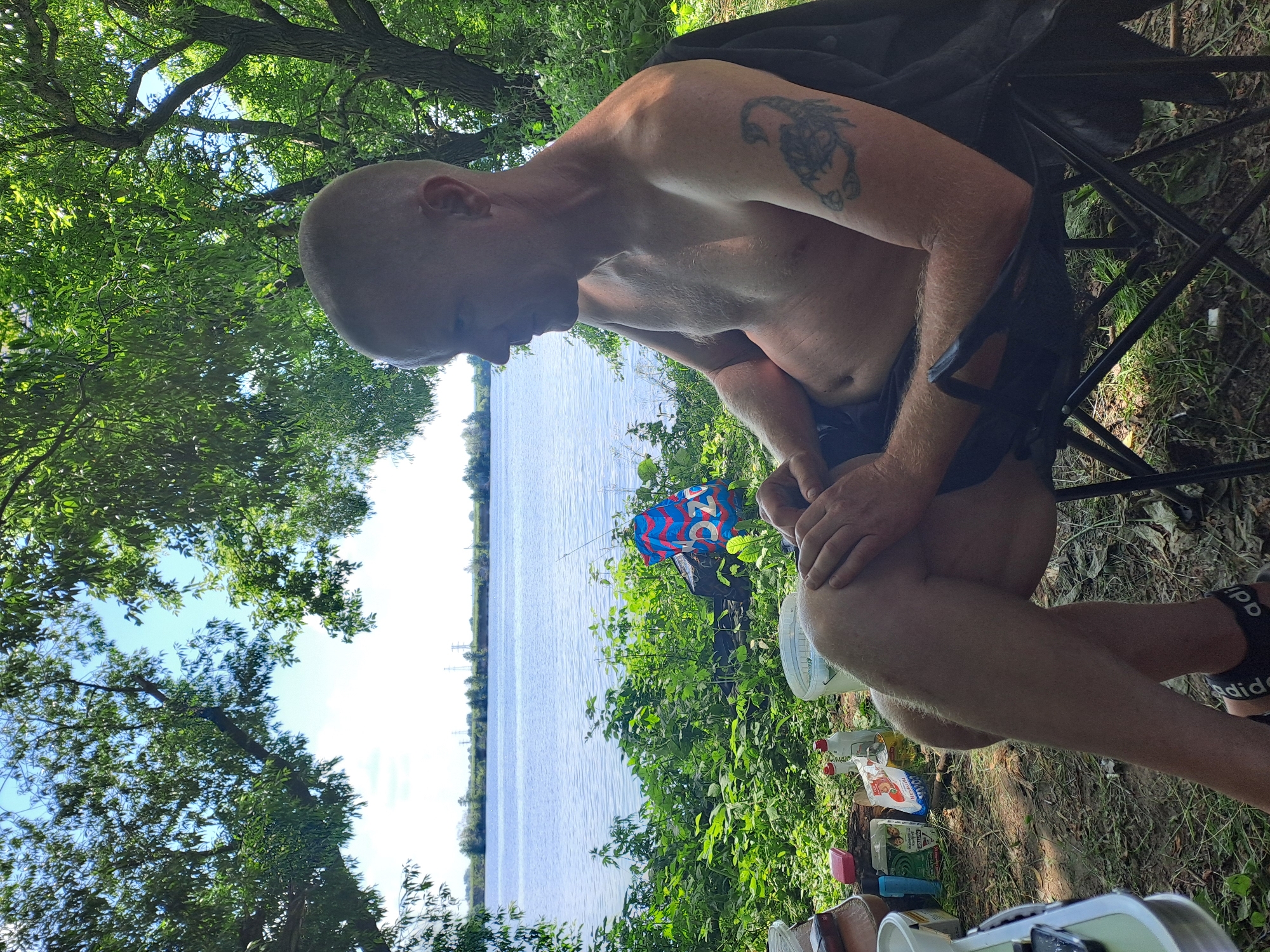 A man with a shaved head, bare-chested, with a scorpion tattoo on his right arm, sits in a folding chair near a body of water, possibly the Oka River in Dedinovo, Russia. He is looking to the right, with his legs crossed and hands resting on his lap. A fishing rod leans against the vegetation behind him, and a colorful bag lies on the ground. Various containers and items are scattered on the ground in front of him, suggesting a picnic or camping setup. The background features lush green foliage and trees framing a tranquil lake or river under a clear blue sky.