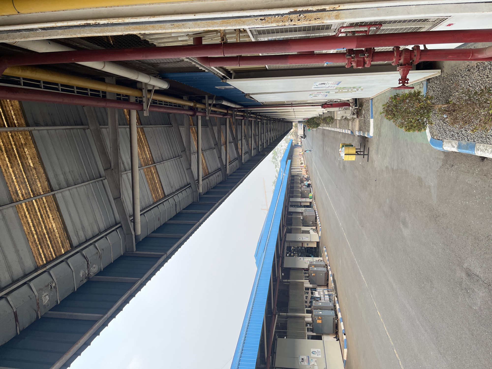 The image depicts an outdoor scene at an industrial facility, likely a warehouse or factory, in Pelang, Indonesia. The perspective is from the side of a long, covered walkway, looking down a paved road. The walkway has a corrugated metal roof, with some sections appearing weathered and rusty. Blue metal siding runs along the base of the walkway structure.
Along the left side of the road, there are several large, grey industrial bins and what appear to be air conditioning units. The road itself is paved with asphalt and has a yellow line painted down the center. The right side of the road is bordered by a low concrete curb with blue and white markings. Beyond the curb, there are some small, manicured bushes planted in gravel.
The building facade on the right side is a light color, with prominent red and yellow pipes running vertically. There are also sections of white siding and what appears to be a metal grate. A red fire hydrant with a visible wheel valve is mounted on one of the red pipes.
In the distance, more buildings with blue roofs can be seen, as well as what looks like a construction crane. A few individuals are visible walking or standing in the distance, and a bicycle is parked near one of the buildings. The sky is overcast, giving the scene a muted, slightly hazy appearance. The overall impression is one of an active but orderly industrial environment.