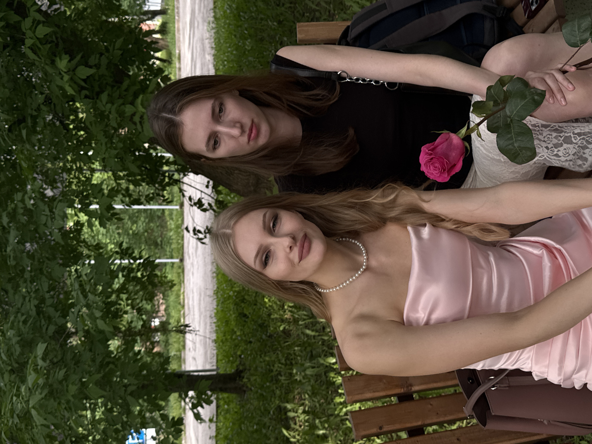 Two young women are sitting on a bench in a park. One woman has long brown hair and is wearing a black shirt. The other woman has long blonde hair and is wearing a pink dress and a pearl necklace. They are holding a pink rose. A bag is on the bench. The setting appears to be a park, with trees and a path visible. There is street furniture visible in the background and the location of this photo may be the city of Perm, Russia.