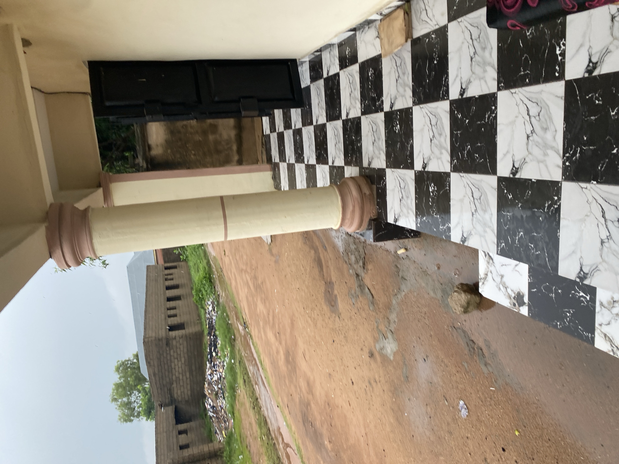 This image depicts an exterior view of a building with a tiled walkway in the foreground, situated in Jalingo, Nigeria. The walkway features a checkered pattern of black and white marble-effect tiles. To the left of the walkway, a dirt path is visible, showing signs of recent rain with puddles and mud. Further to the left, a pile of assorted trash and debris is accumulated near a brick building with multiple openings. A large tree stands behind the brick structure. The building in the foreground has a covered porch supported by a large, fluted column. A dark metal door is visible slightly ajar, leading into the building. The overall scene suggests an unfinished or developing area, with elements of disarray alongside newly laid tiling.