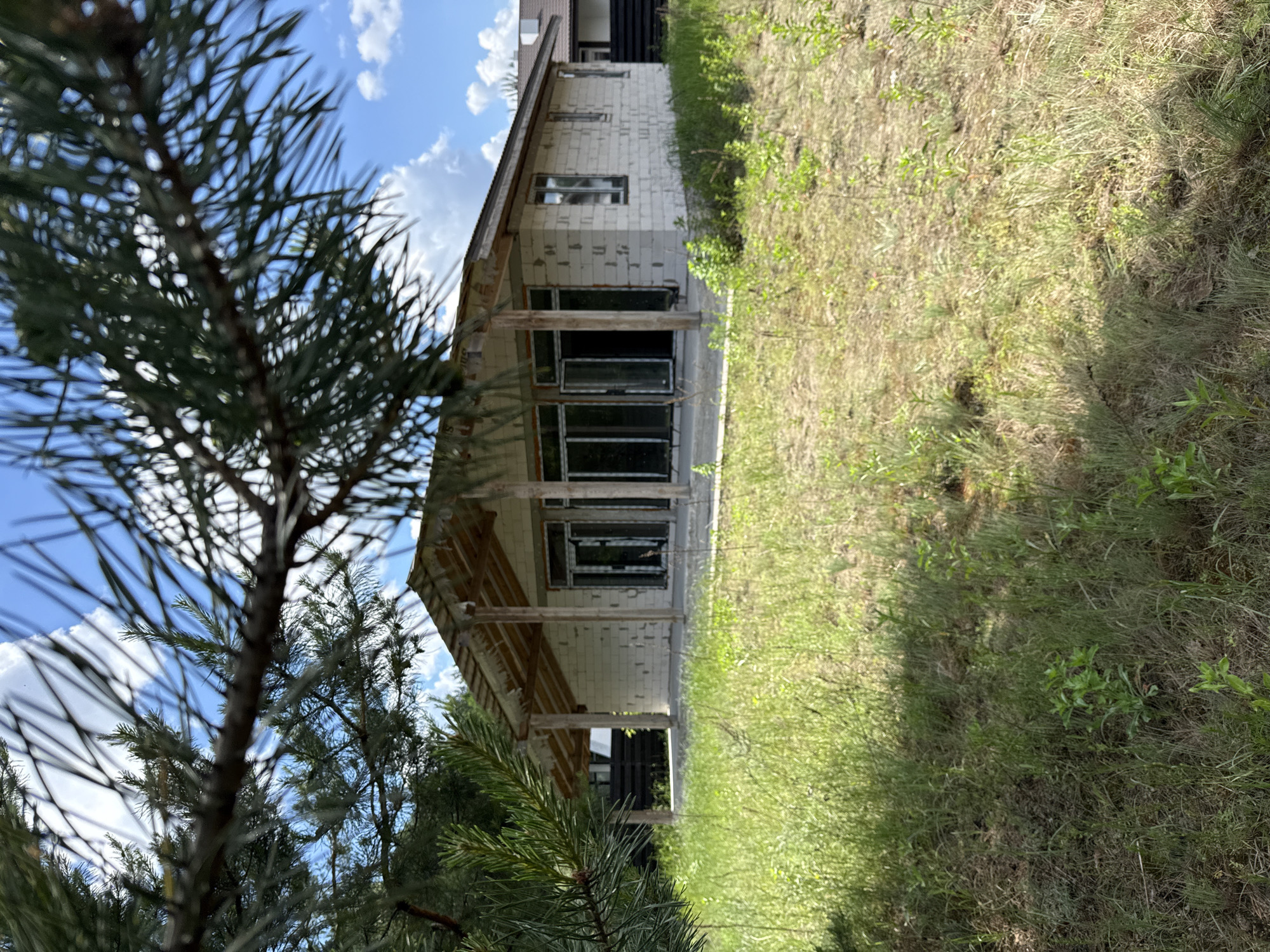 A single-story house under construction is viewed through pine branches in Khmilna, Ukraine. The house is made of cinder blocks and has large glass doors and windows. A covered porch extends across the front of the structure, supported by wooden posts. The surrounding area is overgrown with tall grass and weeds, and the sky above is blue with scattered white clouds.