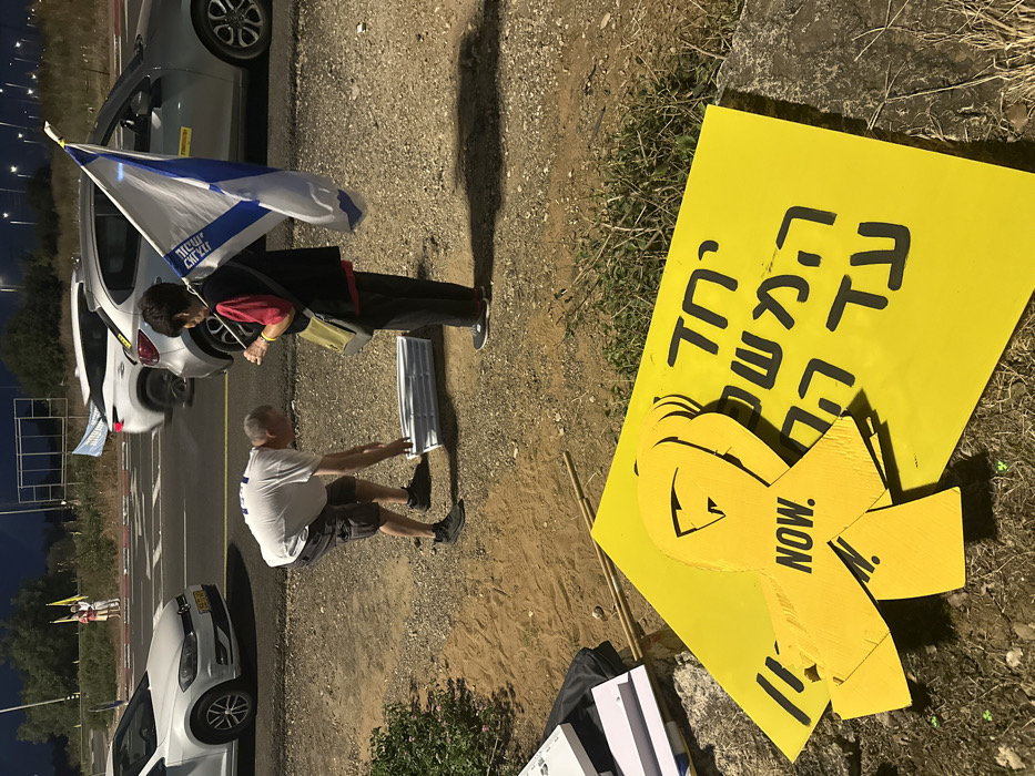 The image captures a nighttime scene on a dirt roadside shoulder in Mishmar Hasharon, Israel. In the foreground, multiple yellow signs are visible on the ground, along with several yellow ribbon-shaped cutouts. One prominent yellow ribbon displays "NOW." in black text, while a larger yellow sign features Hebrew script. A man, wearing a white t-shirt, dark shorts, and dark athletic shoes, is bent over, organizing or collecting a stack of white rectangular items from the ground. Adjacent to him, an older woman, dressed in a dark vest over a red top, dark pants, and dark athletic shoes, stands holding an Israeli flag upright on a pole. The flag is white and blue with a central Star of David and partially visible Hebrew text. In the background, a paved two-lane road carries vehicular traffic, including a white Volkswagen Golf (Israeli license plate 59:267:03) and a silver SUV. One vehicle displays a yellow "Bring Them Home" sticker on its rear window, and another has a light blue banner protruding. Distant figures holding yellow flags are faintly visible further down the road. The immediate ground consists of packed dirt, gravel, and sparse green vegetation, illuminated by artificial light sources, likely streetlights.