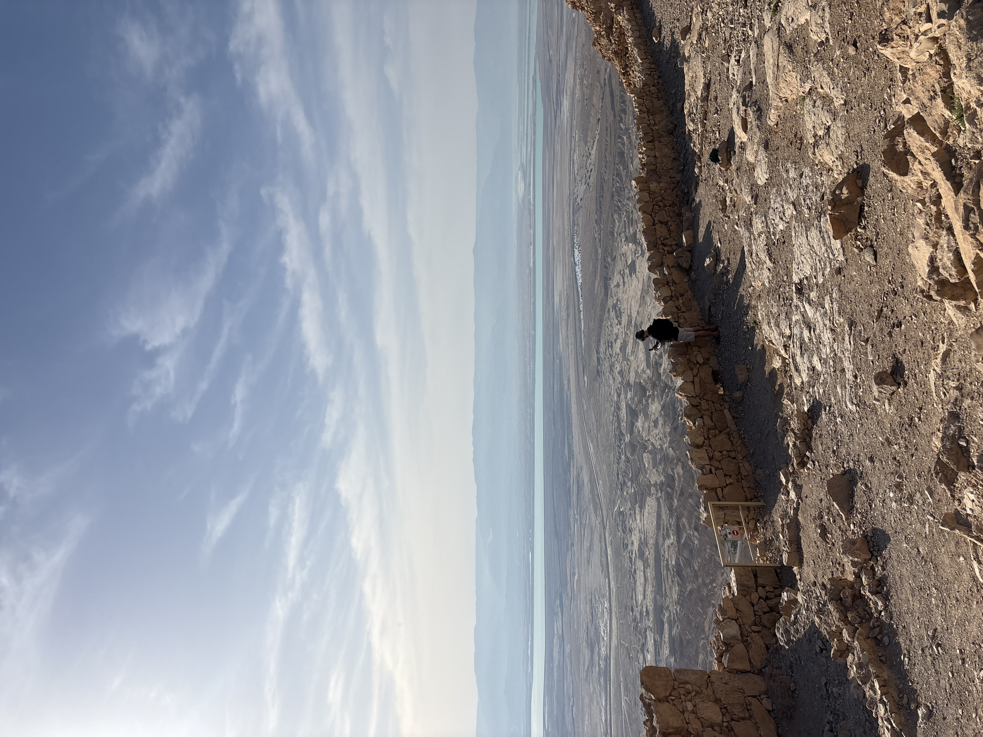 The image shows a landscape in Tamar Regional Council, Israel, with a clear, partly cloudy sky dominating the upper left. A vast, dry, desert-like landscape with a body of water runs horizontally through the center. A man is standing near a stone retaining wall on the right side. The terrain is rocky and uneven, with a security barrier or railing present.