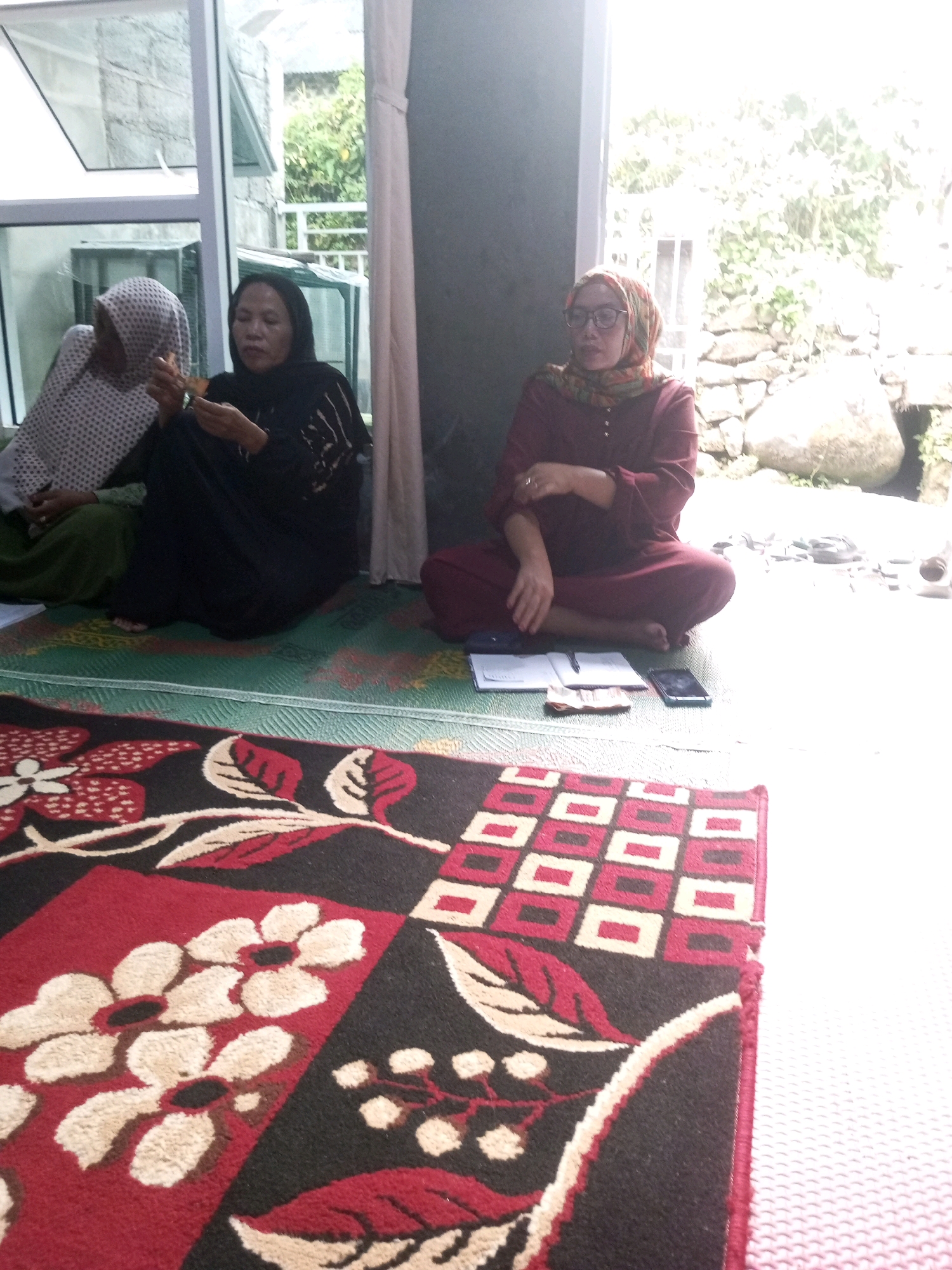 Three women are sitting on a floor mat in front of an open glass door, with a natural stone wall and foliage visible outside. One woman on the left wears a light-colored patterned headscarf and a green garment, with her hands near her face. In the center, a woman in a black abaya holds something in her hands. To the right, a woman with glasses and a red headscarf, wearing a maroon outfit, is seated with her arms crossed. In front of the seated women are papers, a phone, and some cash. A patterned rug with floral and geometric designs is in the foreground. The scene appears to be an outdoor or semi-outdoor setting in Andongsili, Indonesia.