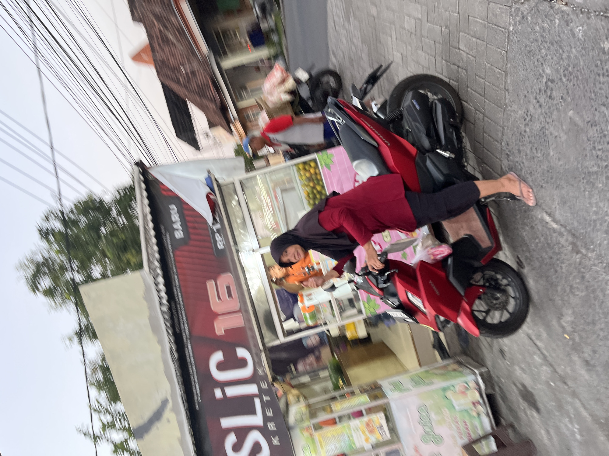 A slightly blurry, medium-shot, eye-level outdoor image captures a woman in a red hijab and long-sleeved red shirt, black pants, and flip-flops. She is positioned on a bright red motorcycle, facing the camera and slightly to the left, with her right hand raised. The motorcycle is parked on a paved surface next to a stall. Behind her, a stall is visible with a large sign displaying "SLIC 16 KRETEK" in white lettering with a red outline. The stall also features a white flag and displays various products and signage. In the background, a man in a grey and red shirt and blue cap is visible near another motorcycle loaded with goods. The scene appears to be a street in Karangtanjung, Indonesia, with buildings and power lines overhead.