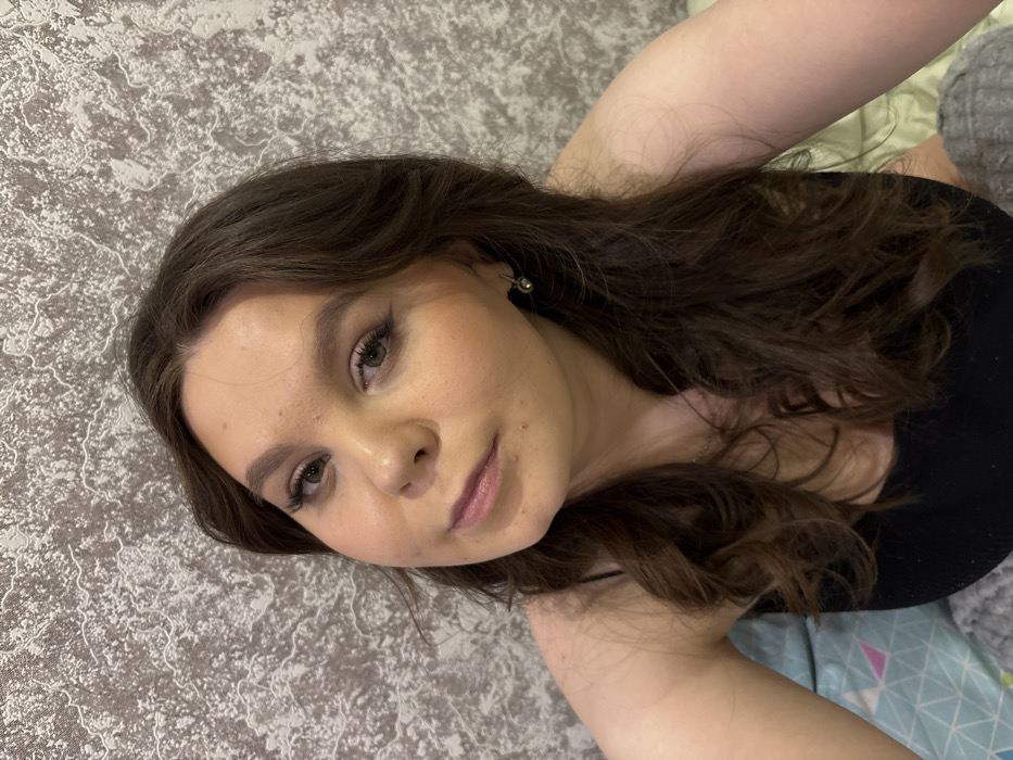 A young woman with light skin and brown hair poses for a selfie. She is tilted slightly with her head angled towards the camera. Her dark brown hair is wavy and falls around her shoulders. She wears subtle makeup, enhancing her eyes with eyeliner and mascara and wearing nude lipstick. A small, silver earring is visible. She is wearing a black top. The background features a patterned rug or wallpaper.
