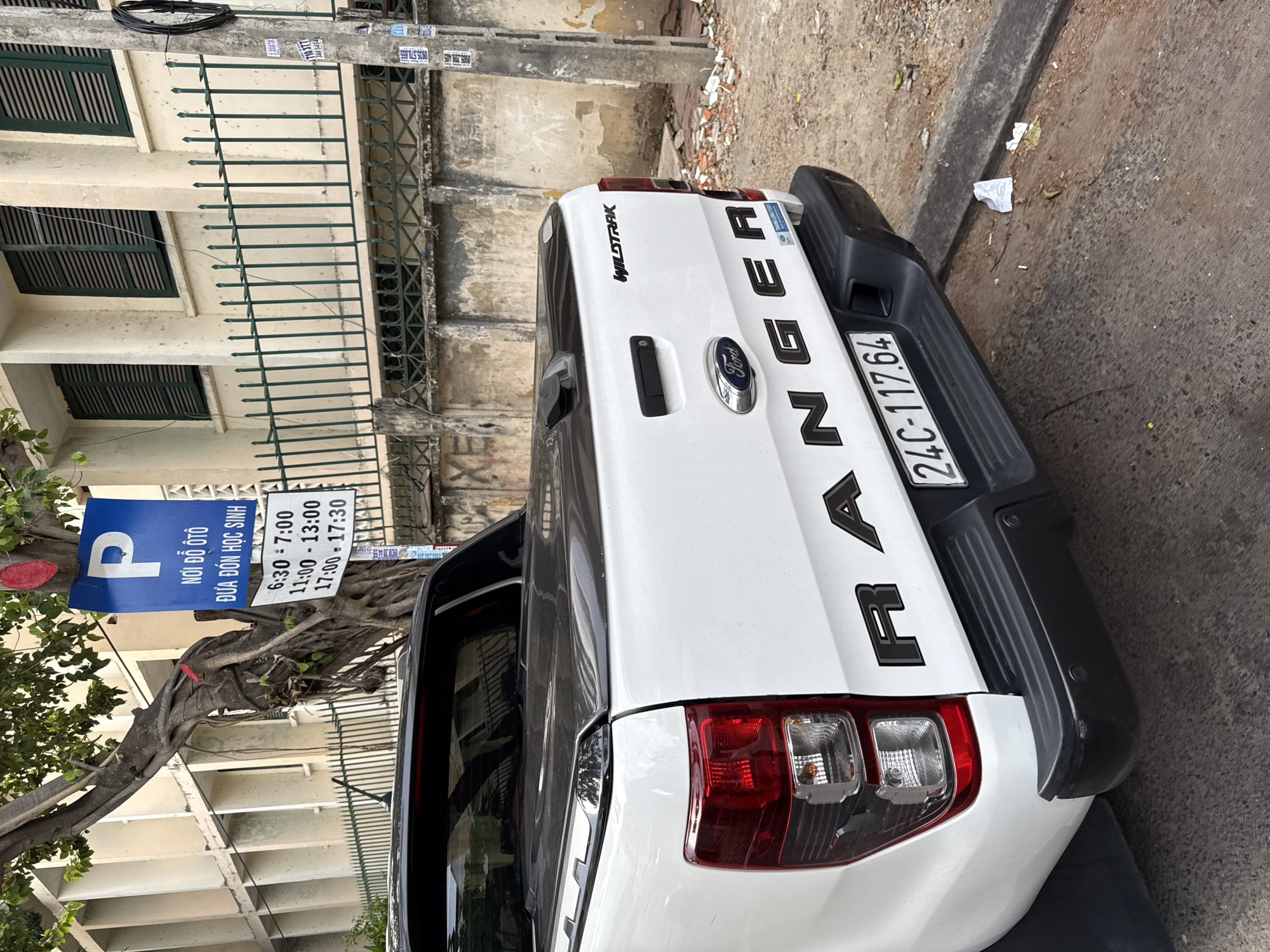 The image depicts the rear passenger side of a white Ford Ranger pickup truck parked on a city street in Nha Trang, Vietnam. The truck has a black tonneau cover over the bed and the "RANGER" and "WILDTRAK" badging is visible. The license plate reads "24C-117.64".
In the background, a building with beige walls and green shutters stands behind a green metal fence. A utility pole is situated to the right of the fence, adorned with various stickers and wires. A tree with gnarled roots grows in front of the building and the fence, partially obscuring a blue parking sign. The sign features a white "P" symbol and Vietnamese text that translates to "NO OTO" (car parking) and "DUA DON HOC SINH" (school student pick-up/drop-off), followed by a schedule of restricted times: 6:30-7:00, 11:00-13:00, and 17:00-17:30. The street surface appears to be concrete with some debris, including broken bricks and scattered paper, near the curb. No people are visible in the image.