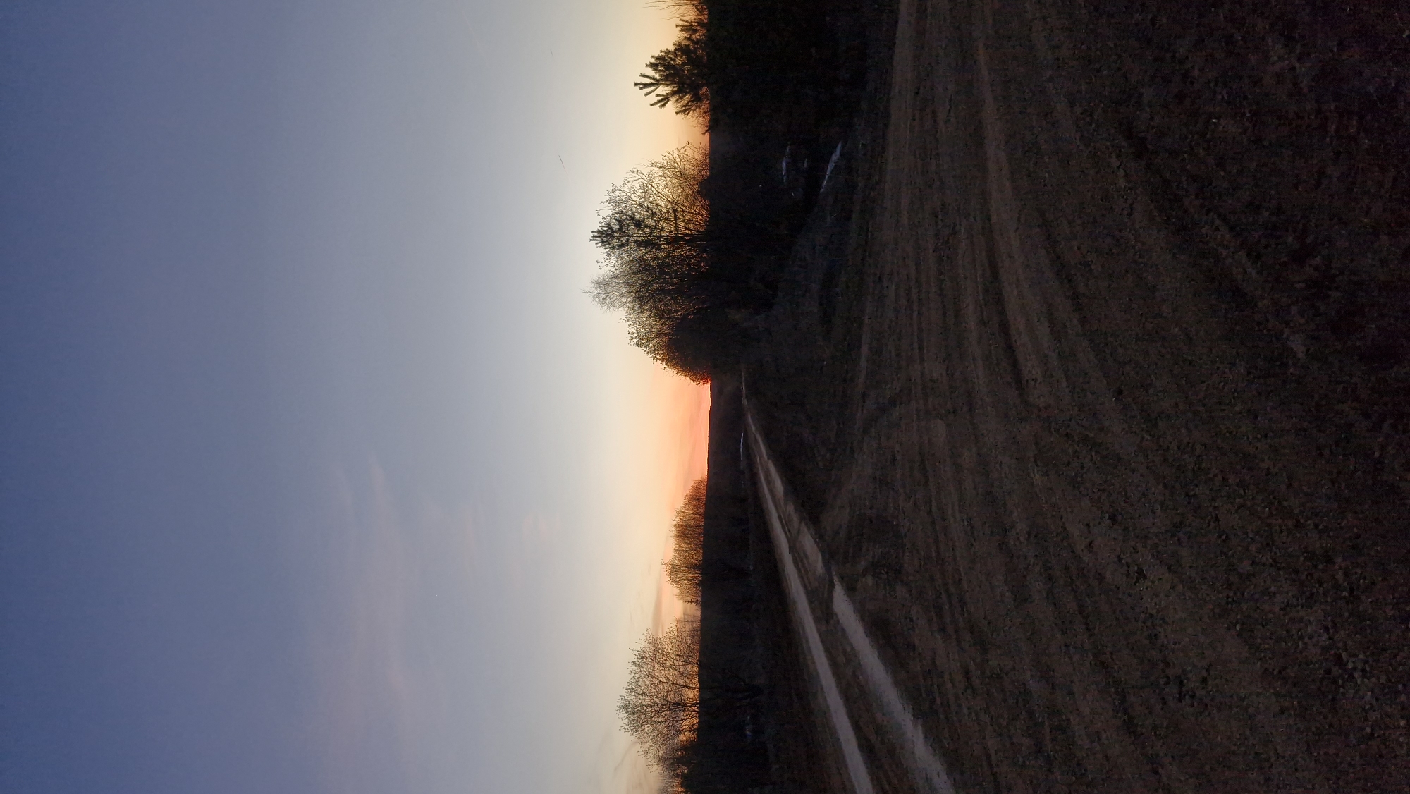 The image shows a dirt road running along the edge of a field or wooded area. The sky is visible, with a gradation from blue to orange, indicating sunset or sunrise. The location is Otchor, Russia. The road is dirt, with tracks suggesting recent vehicle passage. There are no people visible. The vegetation is sparse, possibly dormant due to the season. The overall scene is rural and depicts a quiet, natural environment.