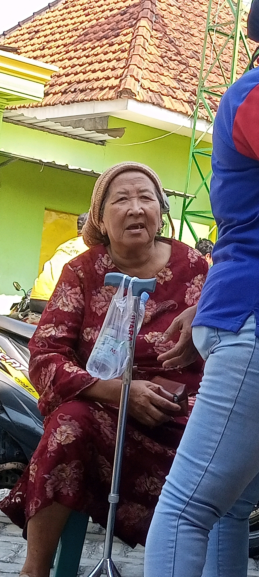 An elderly Indonesian woman wearing a maroon floral dress and a light brown headscarf sits on a motorcycle in Gempolklutuk, Indonesia. She holds a walker with a plastic bag containing a bottle of water. Next to her, a person in a blue shirt and light blue jeans stands, with their hands near the woman. In the background, a green building with a terracotta tiled roof is visible, along with other motorcycles.
